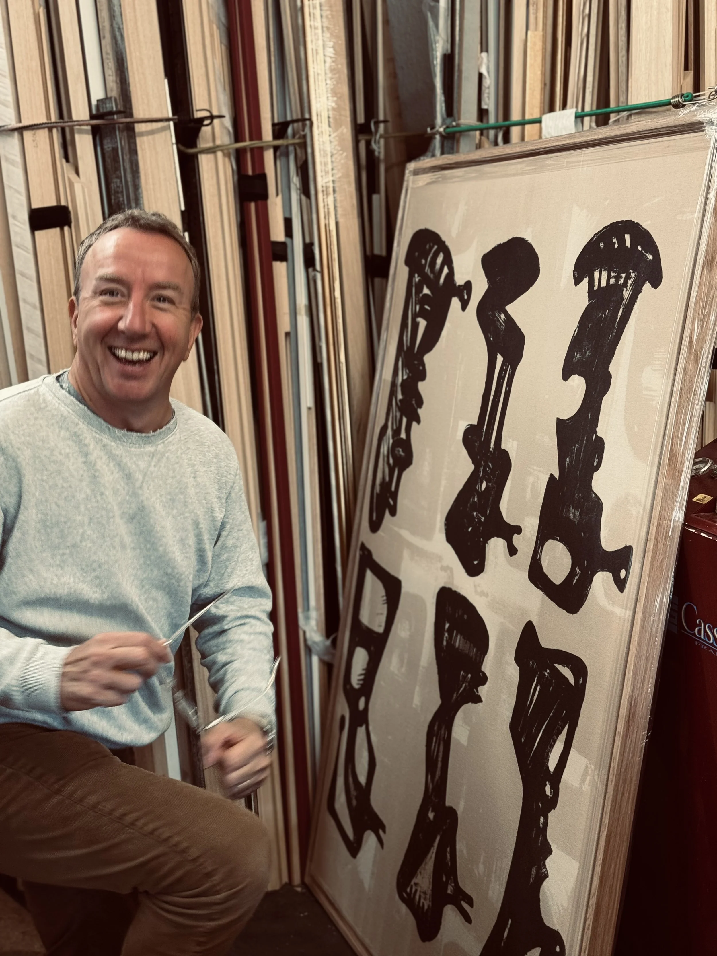 A smiling man in a light gray shirt and brown pants holding a needle and thread stands next to a large black and white animal print artwork in a frame, with wooden framing materials stacked in the background.