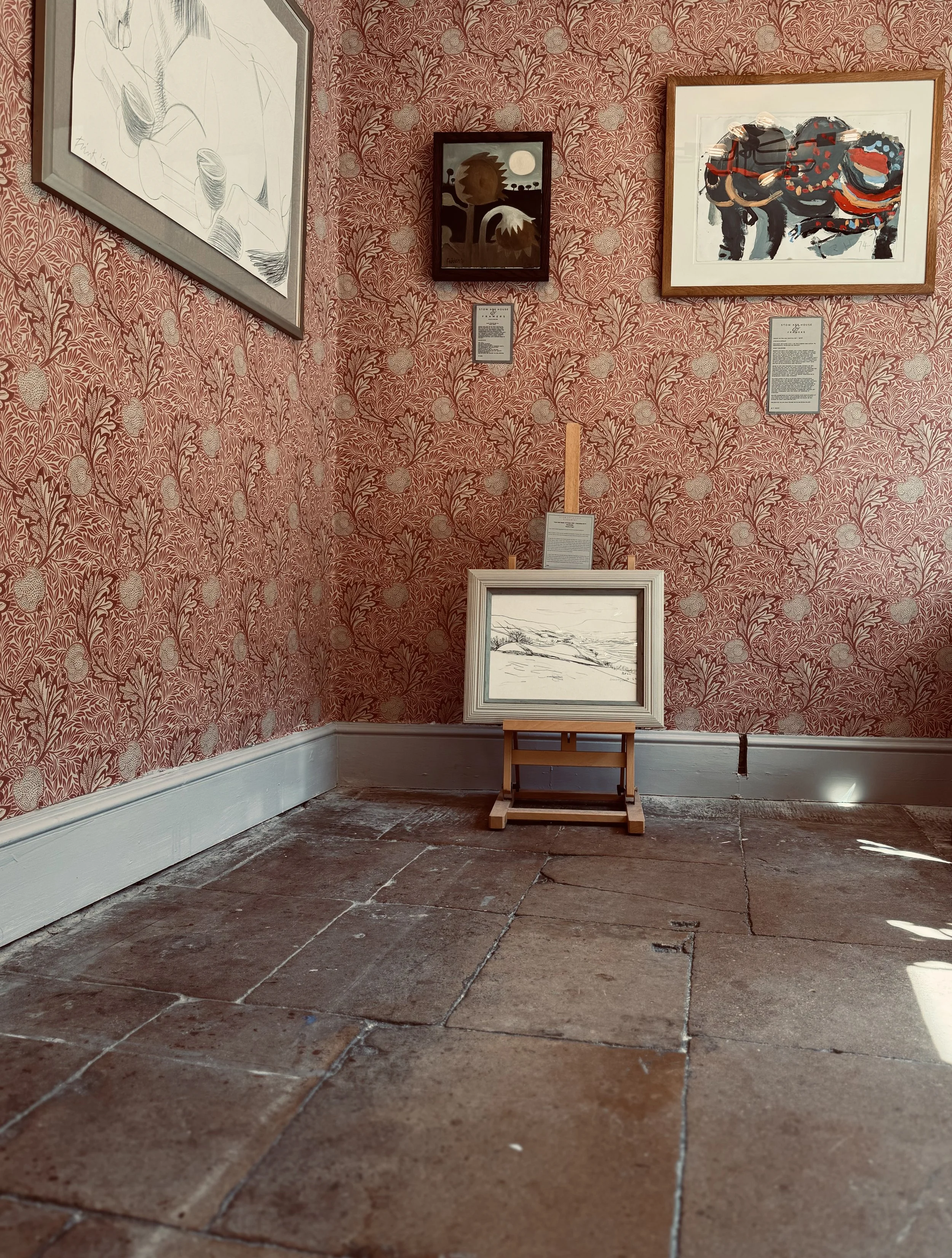 An art gallery scene with a red floral wallpaper, featuring five framed artworks on the wall, a small white framed drawing on the floor, and a wooden easel. The artworks include a sketch of a reclining figure, abstract faces, an animal with a colorfu
