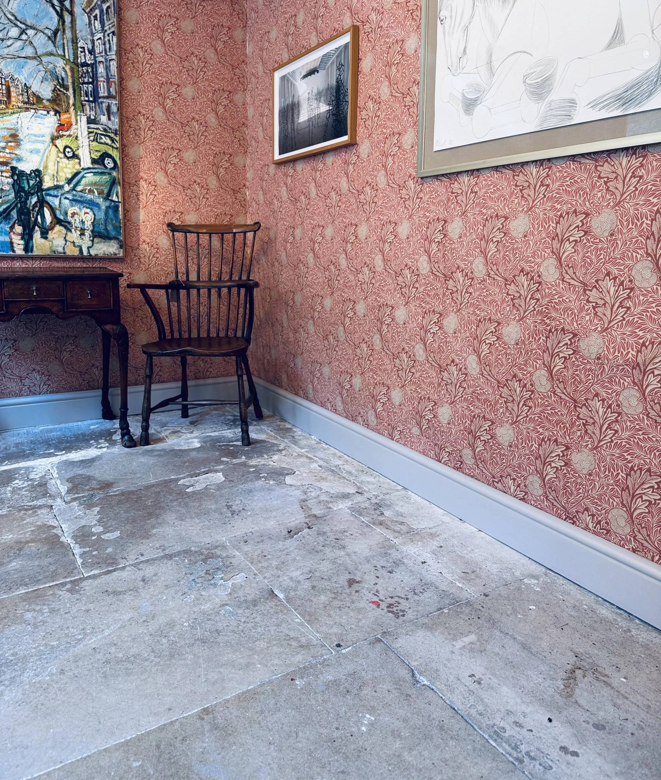 Interior of an art gallery with wallpapered walls, displaying paintings and sketches, including a colorful street scene and a black and white drawing of a horse, with a wooden chair and a small wooden table.