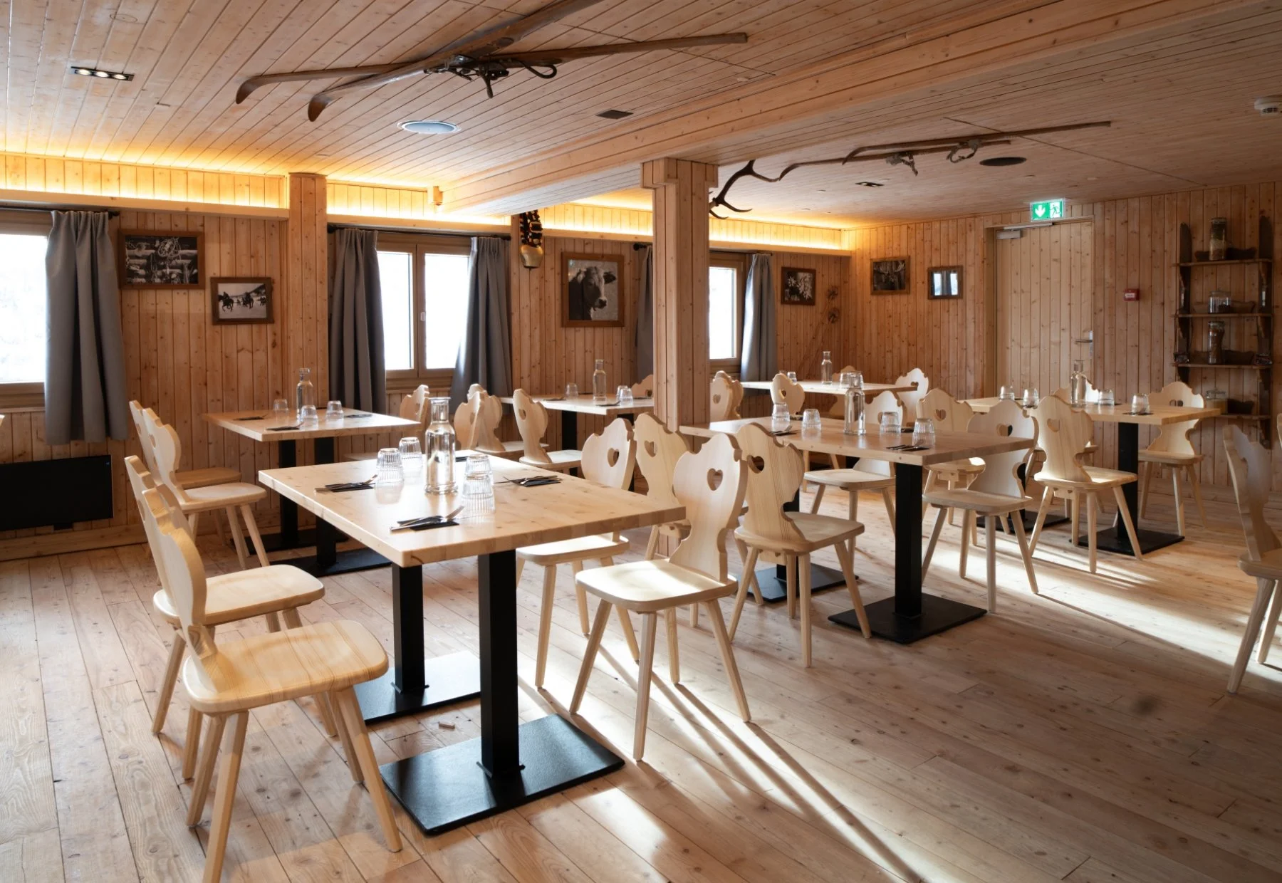 Sala da pranzo in stile rustico con tavoli di legno chiaro, sedie con schienali a cuore, pareti e pavimenti in legno, decorazioni di attrezzi da montagna, fotografie di animali e finestrini con tende grigie.