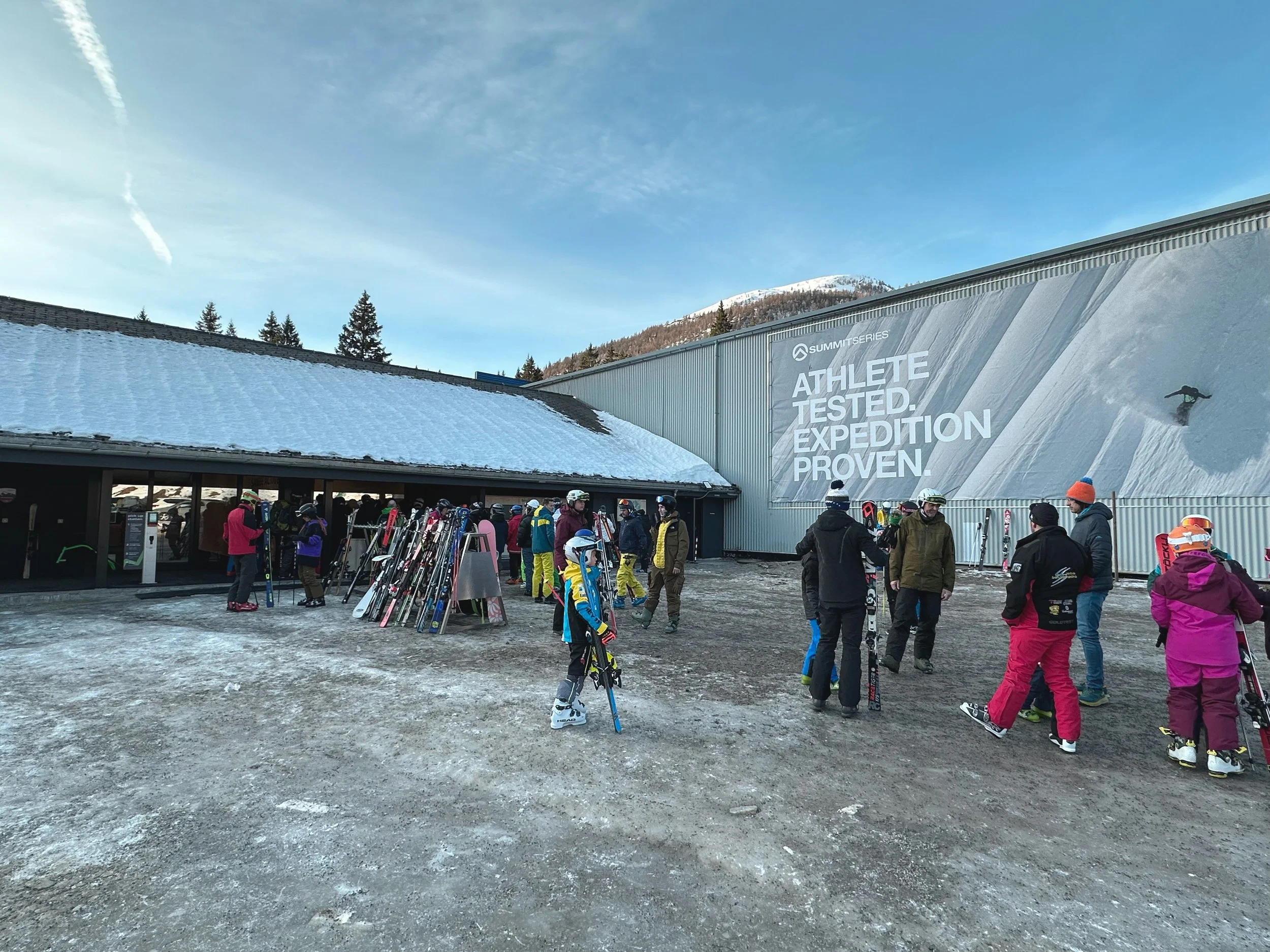 Gruppo di persone con attrezzi da sci davanti a un edificio in montagna, con una pubblicitĂ  sul muro che dice 'ATHLETE TESTED. EXPEDITION PROVEN.' in un resort sciistico.