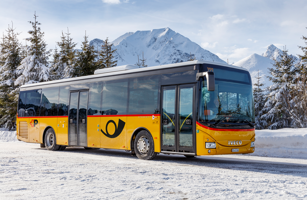 Autobus giallo e nero di IVECO con logo di Poste Italiane, parcheggiato su una strada innevata con montagne e pini ricoperti di neve sullo sfondo.