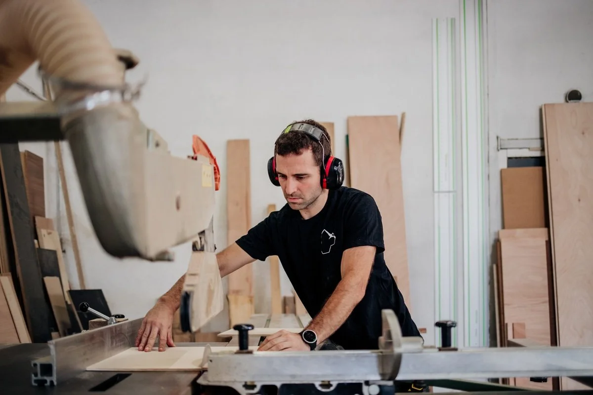 Un homme travaille dans un atelier de menuiserie, portant des protections auditives, avec des planches de bois autour.