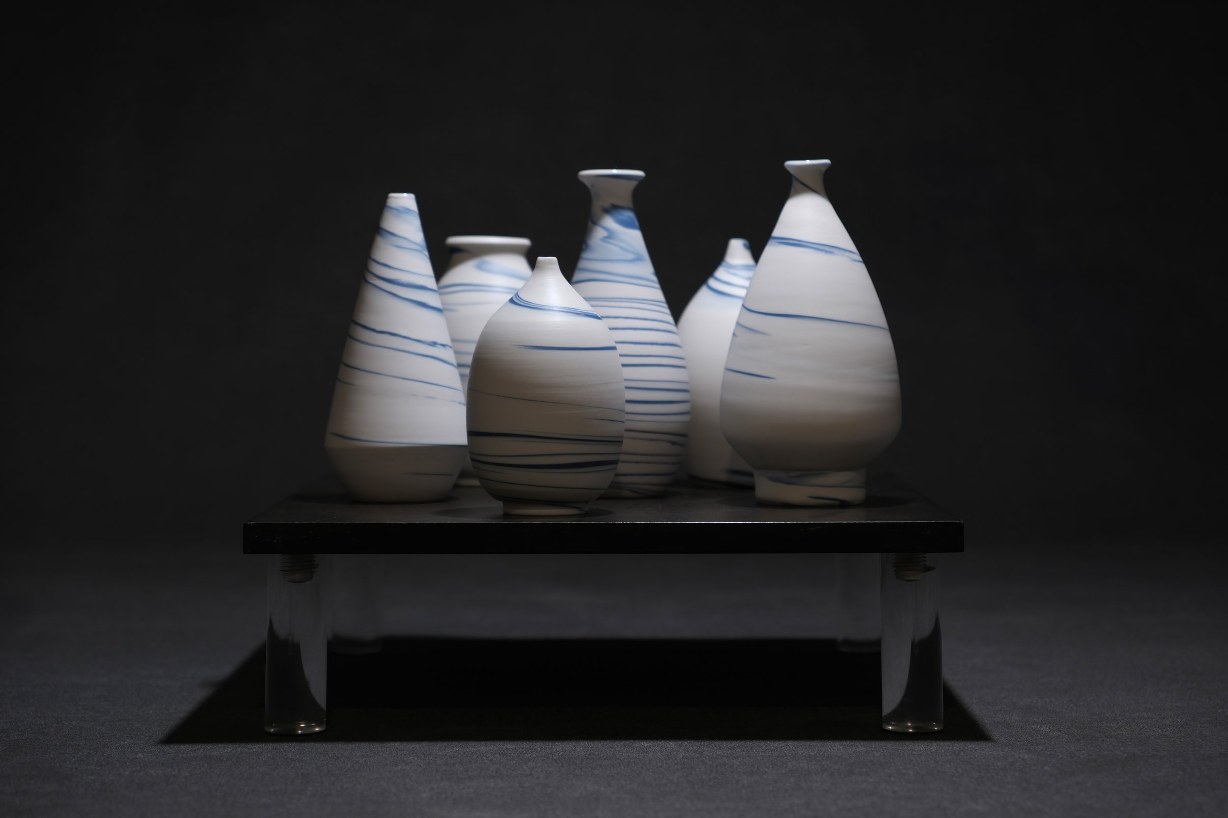 Seven white porcelain vessels with blue swirls on a black surface against a black background.