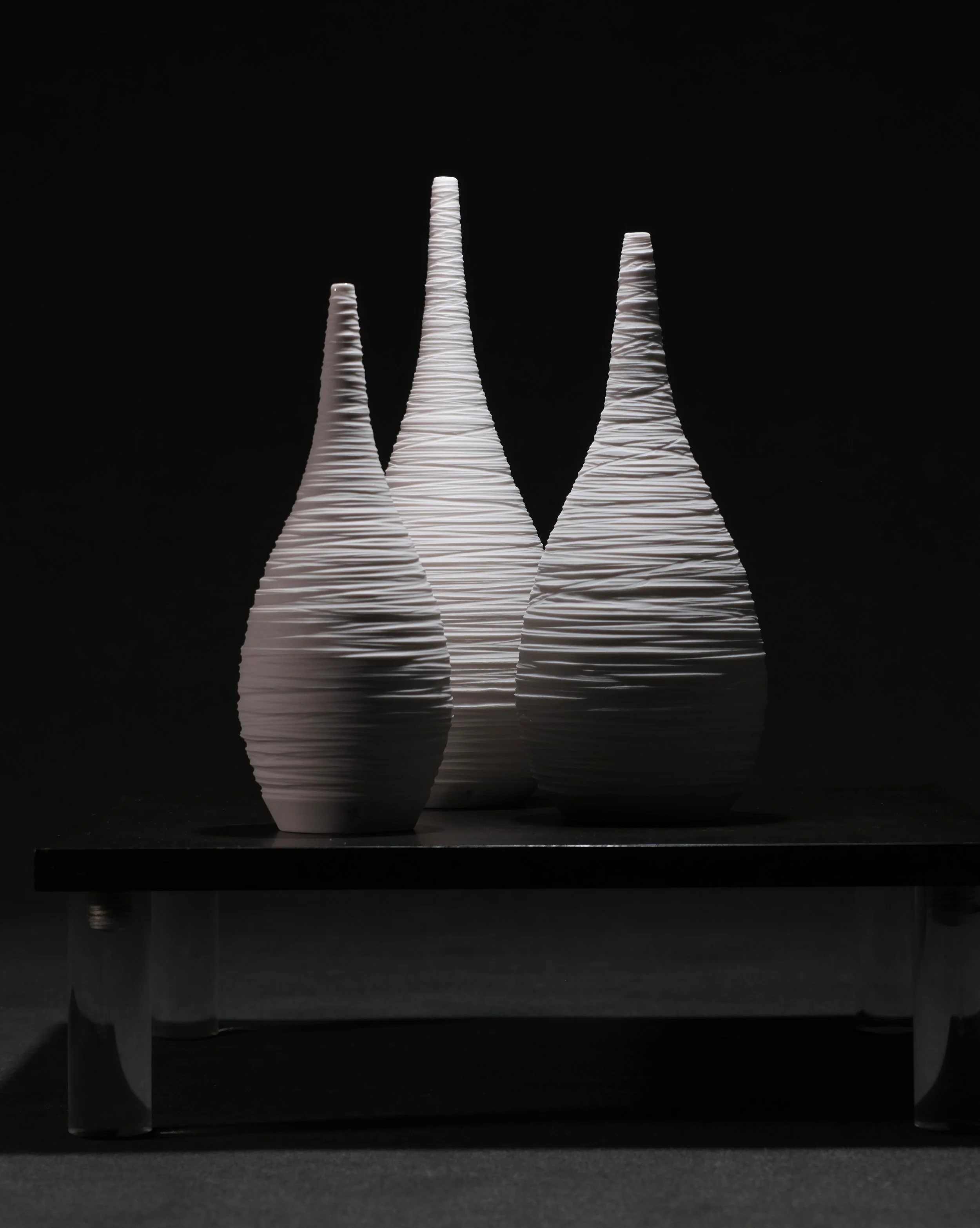 Three tall, cone-shaped ceramic sculptures with ribbed texture, illuminated against a dark background, placed on a black platform.