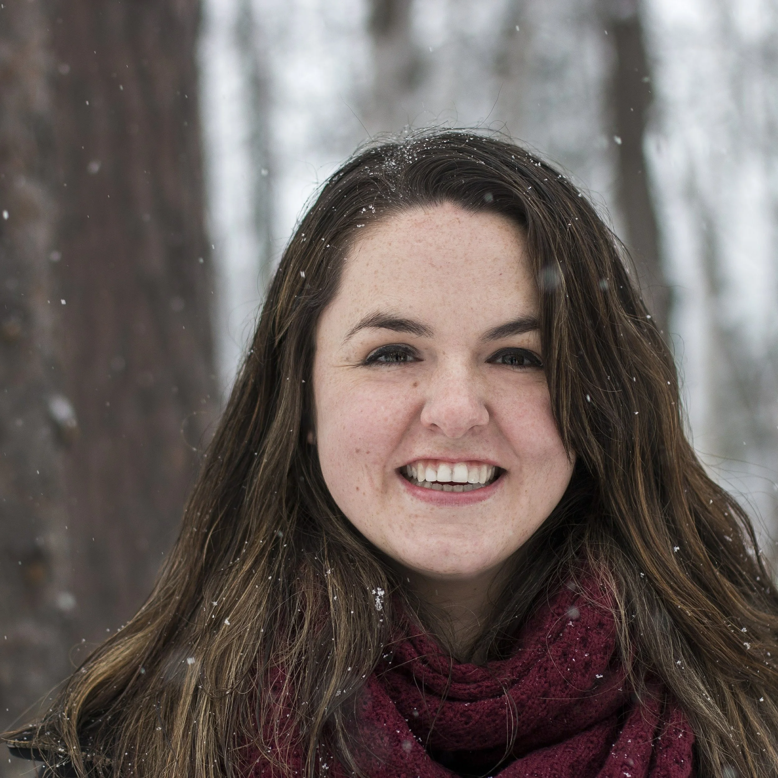 A smiling woman with long brown hair outdoors in a snowy forest.