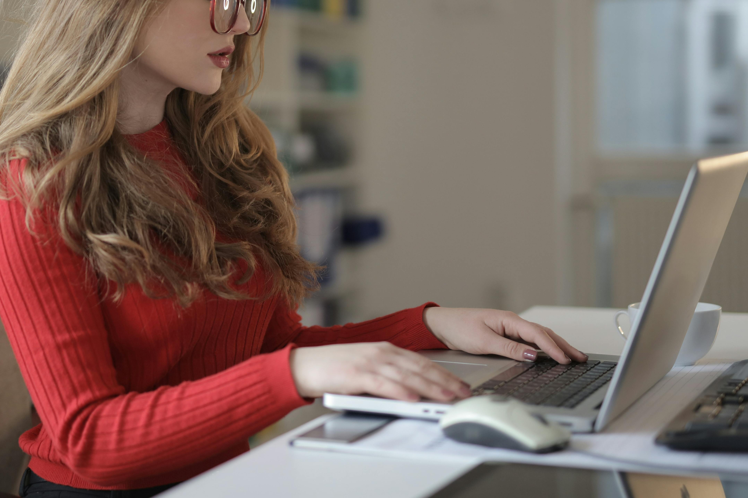 A woman with long red hair, wearing glasses and a red shirt, working on a laptop at a desk in an office setting.