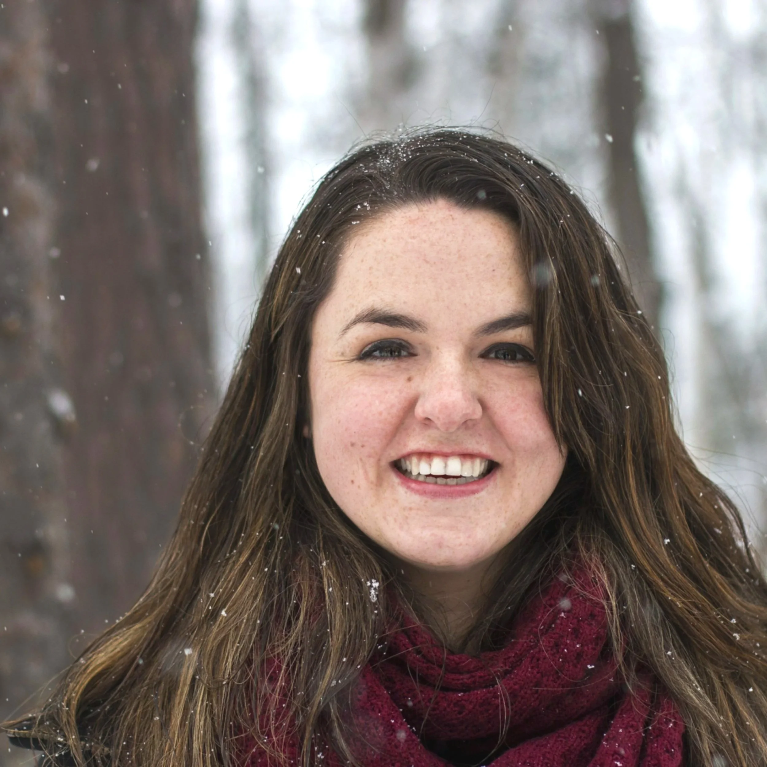 A smiling woman with long brown hair and blue eyes, wearing a burgundy scarf, outdoors in a snowy forest.