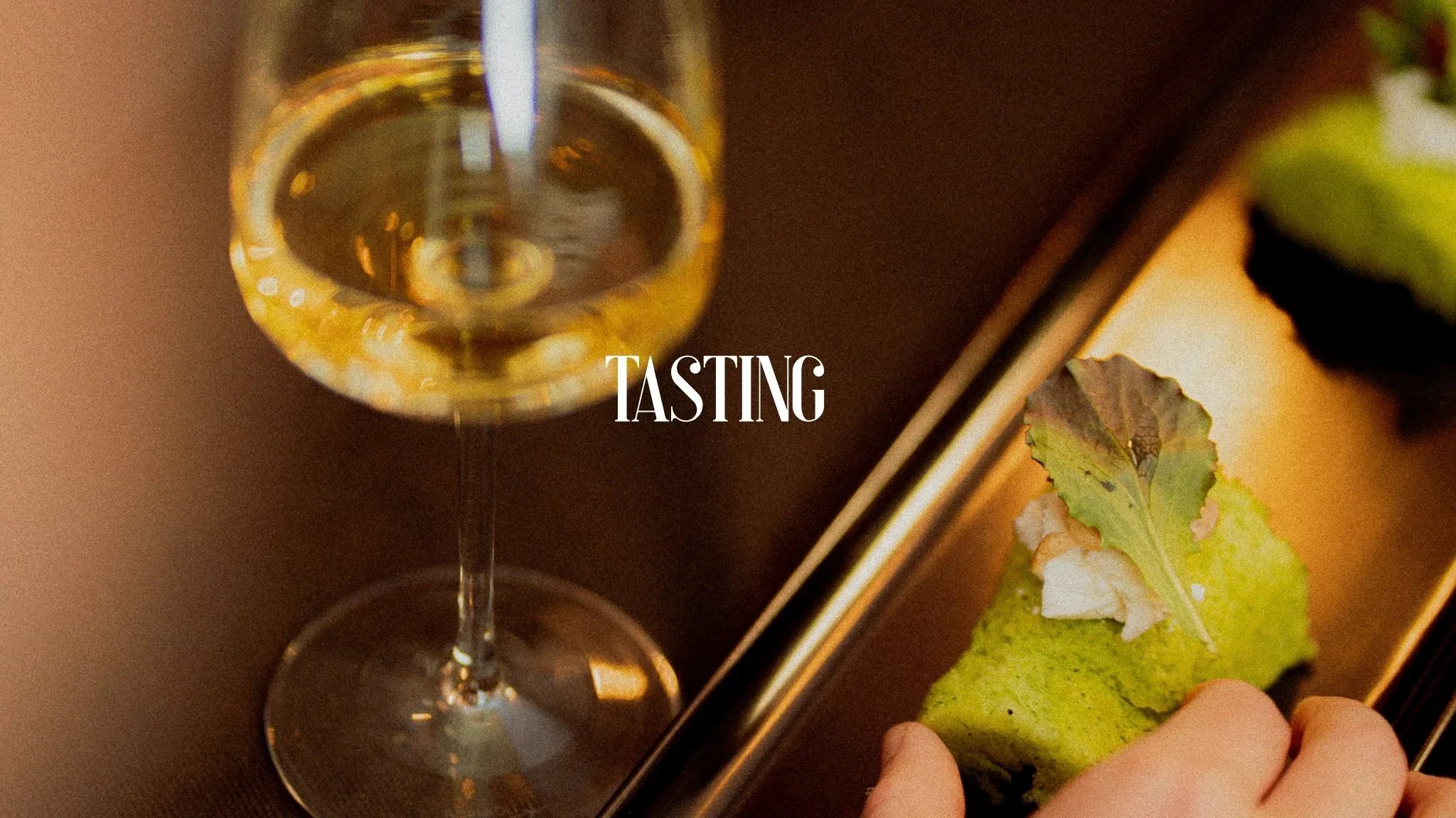 A glass of white wine and a piece of green dessert with a leaf on top, on a reflective surface with the word 'TASTING' in the center.