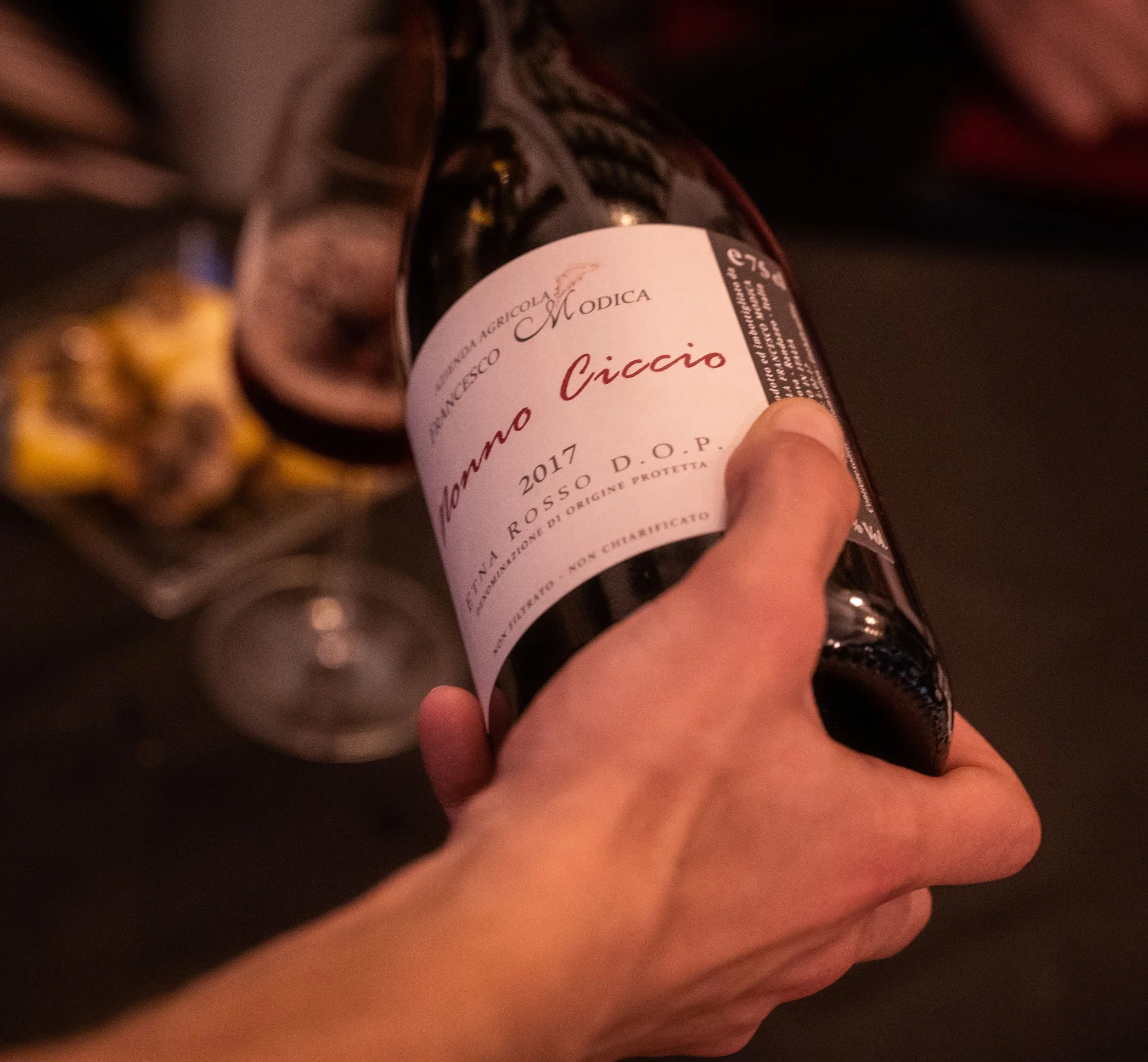 Hand holding a bottle of Mommo Ciccio 2017 red wine with a blurred background of wine glasses and snacks.