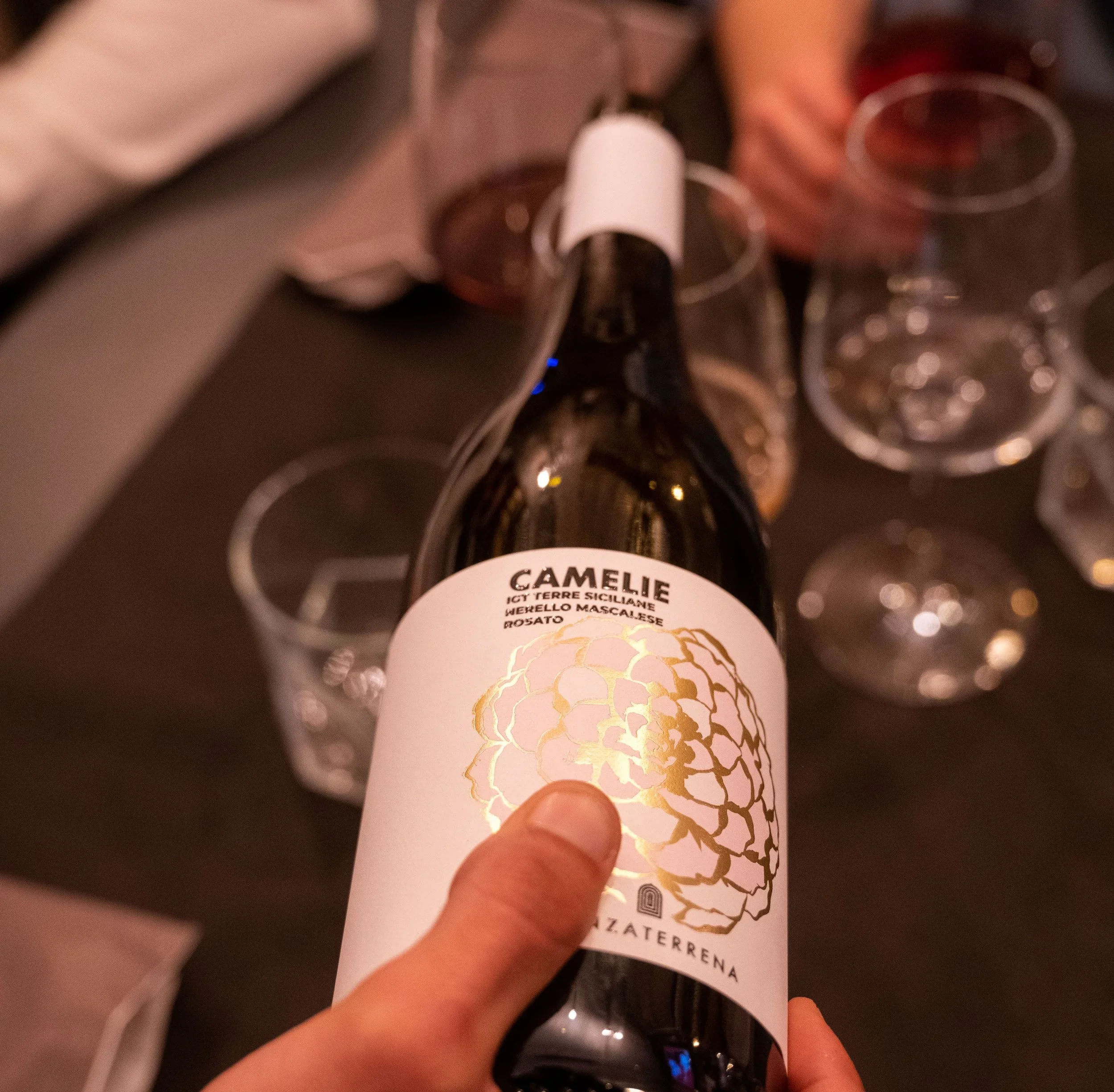 A person holding a bottle of Camelie wine in front of a black table with several empty wine glasses around it.