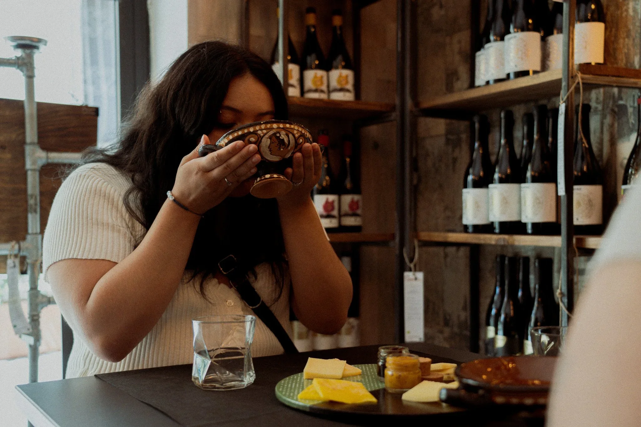 Woman with dark hair tasting cheese in a store, shelves with wine bottles in the background, cheese and jam on the table.