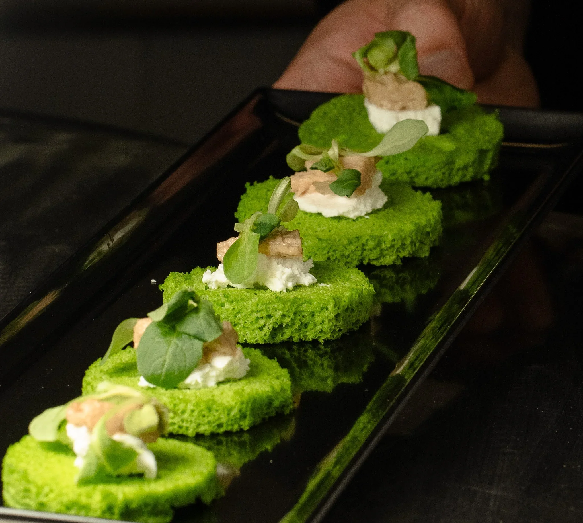 Green savory bread slices topped with cheese, pâté, and microgreens, served on a black rectangular platter.