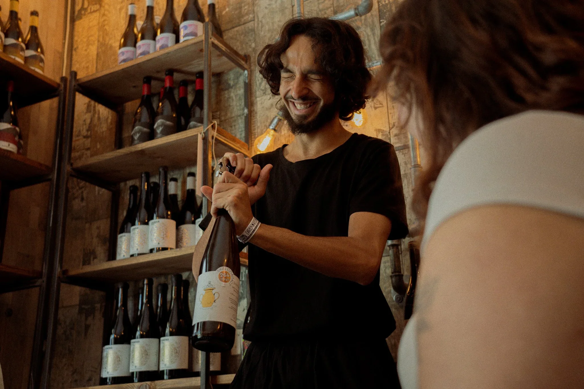 A man with dark, curly hair and a beard smiling and opening a bottle of wine or beverage inside a wine shop. Shelves of bottles are visible behind him, and a woman with brown hair and a white top is partially visible in the foreground.