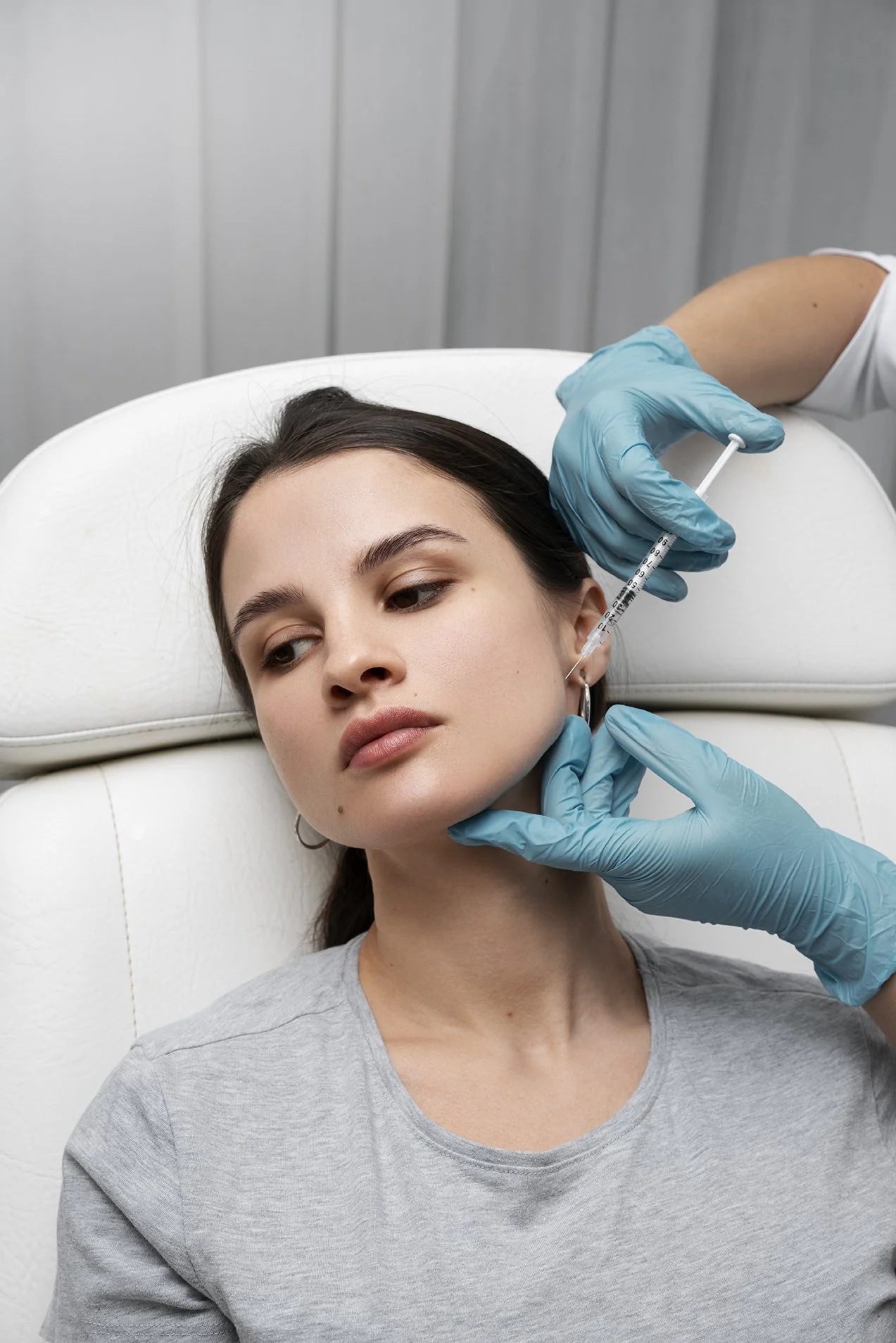 A woman with dark hair lying on a medical chair, receiving a cosmetic injection in her face from a professional wearing blue gloves.