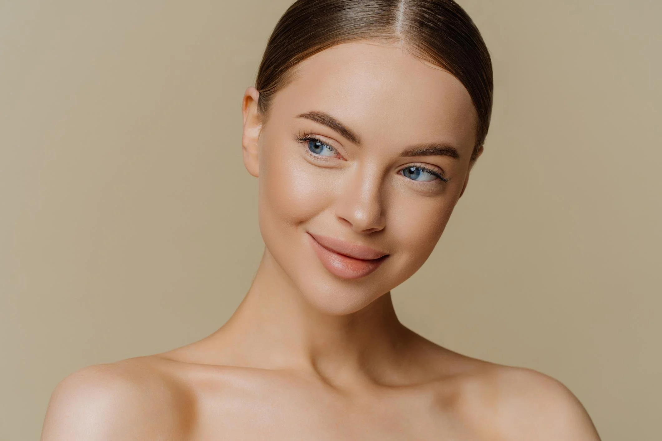 Close-up of a young woman with blue eyes, light skin, and brown hair, smiling softly against a beige background.