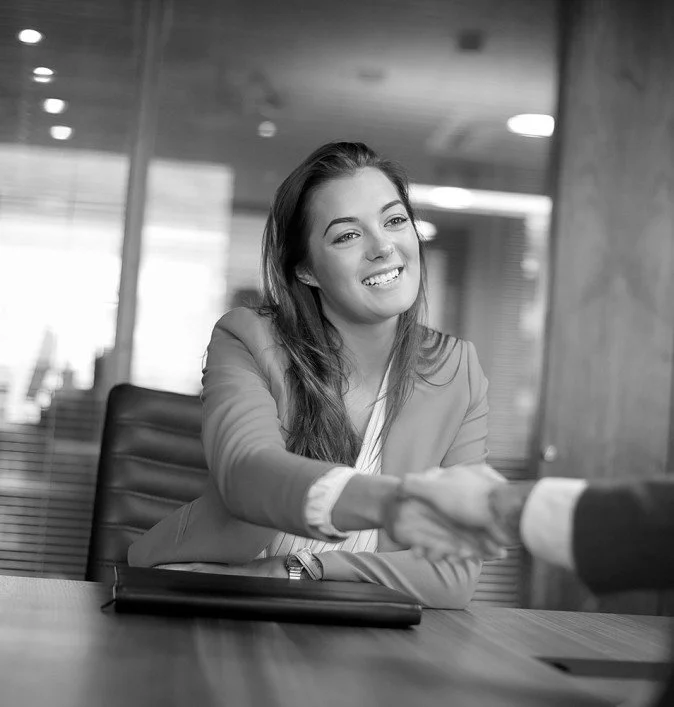 Black and white photo of a woman in business attire smiling and shaking hands with a person off-camera in a modern office setting.