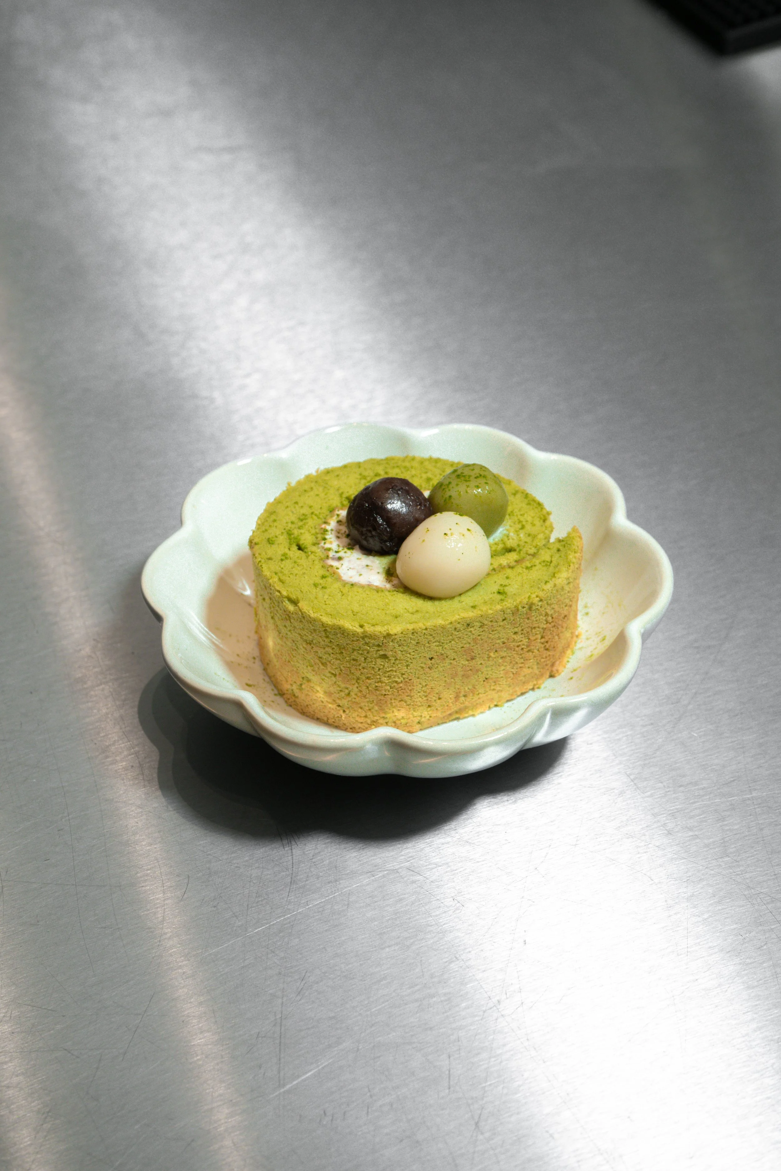 Slice of a matcha redbean rollcake with 2 dangos and one ball of red bean paste in a white dish on a metallic surface.