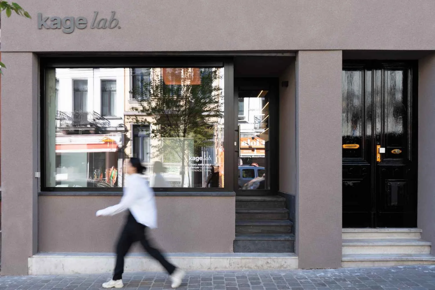 A person walking past a modern storefront with large glass windows and a black door. The store has a sign that reads "kage lab" and is located on a city street with buildings and parked cars reflected in the window.