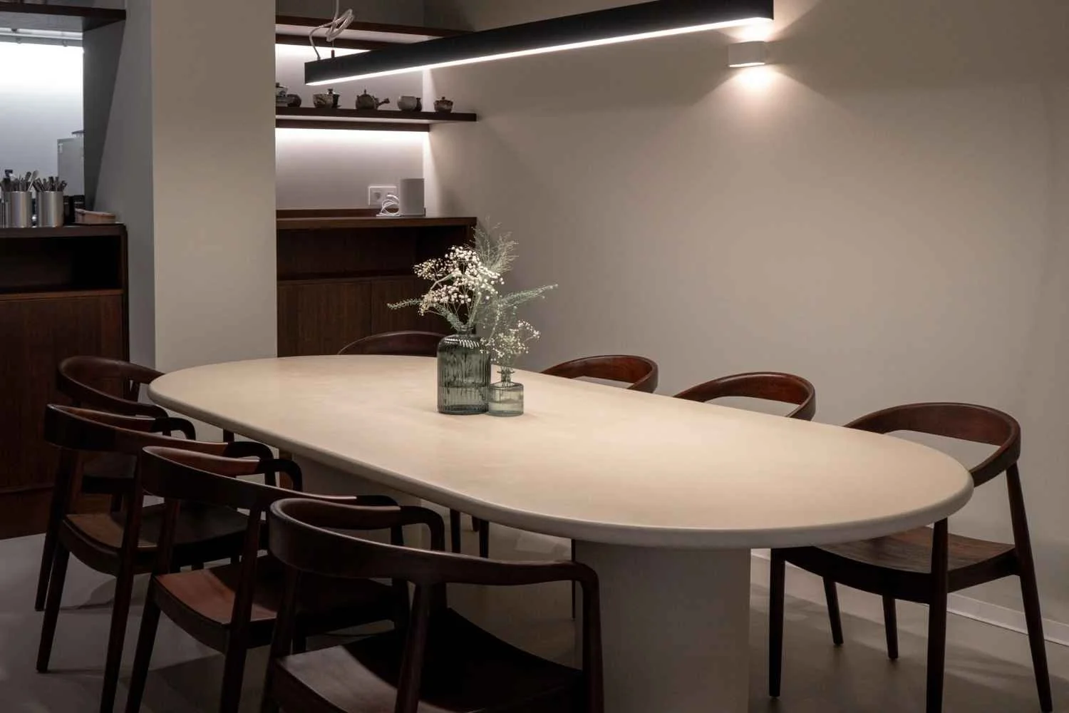 A minimalist sharing oval white table, surrounded by eight wooden chairs, and a small floral centerpiece in a vase. The room has neutral-colored walls, wooden shelves with decorative items, and modern lighting.