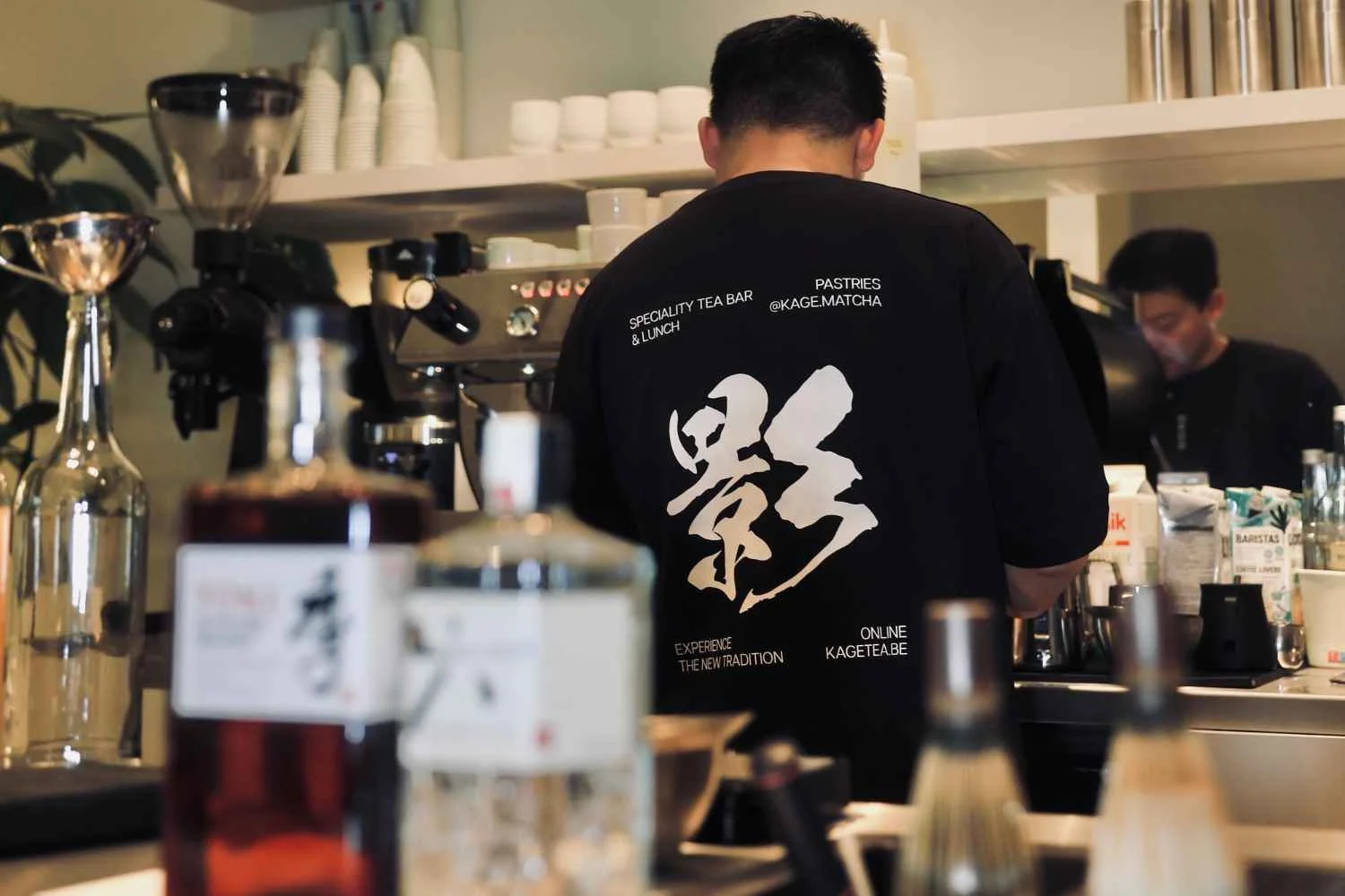 Barman working behind the counter of a tea bar, with coffee-making equipment and bottles in the foreground, and another staff member in the background.