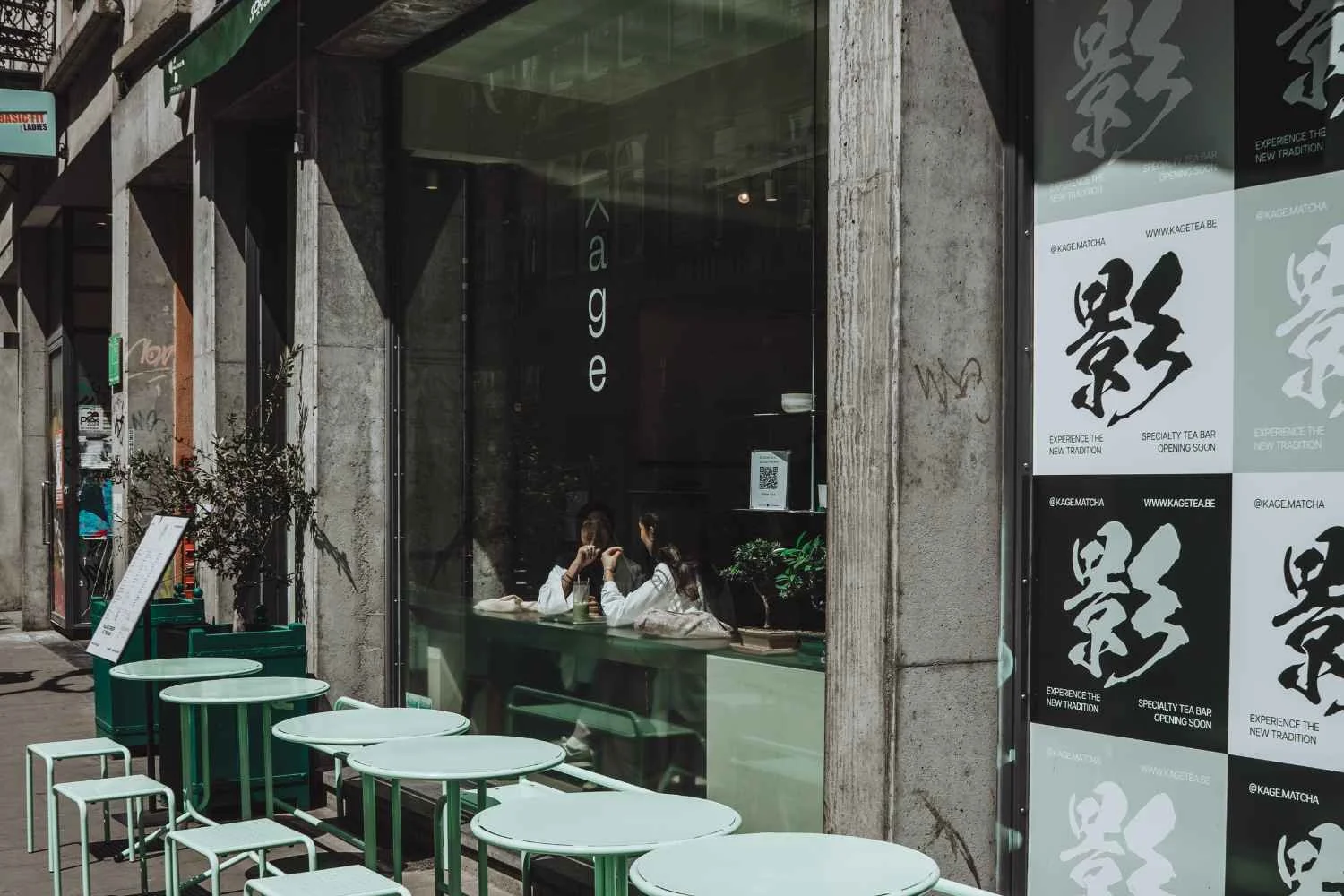 Two women sitting at a table inside a cafe, visible through a large glass window. Several outdoor tables and chairs are outside the cafe on the sidewalk. The building has a concrete exterior, and there are promotional posters on the right side of the window.