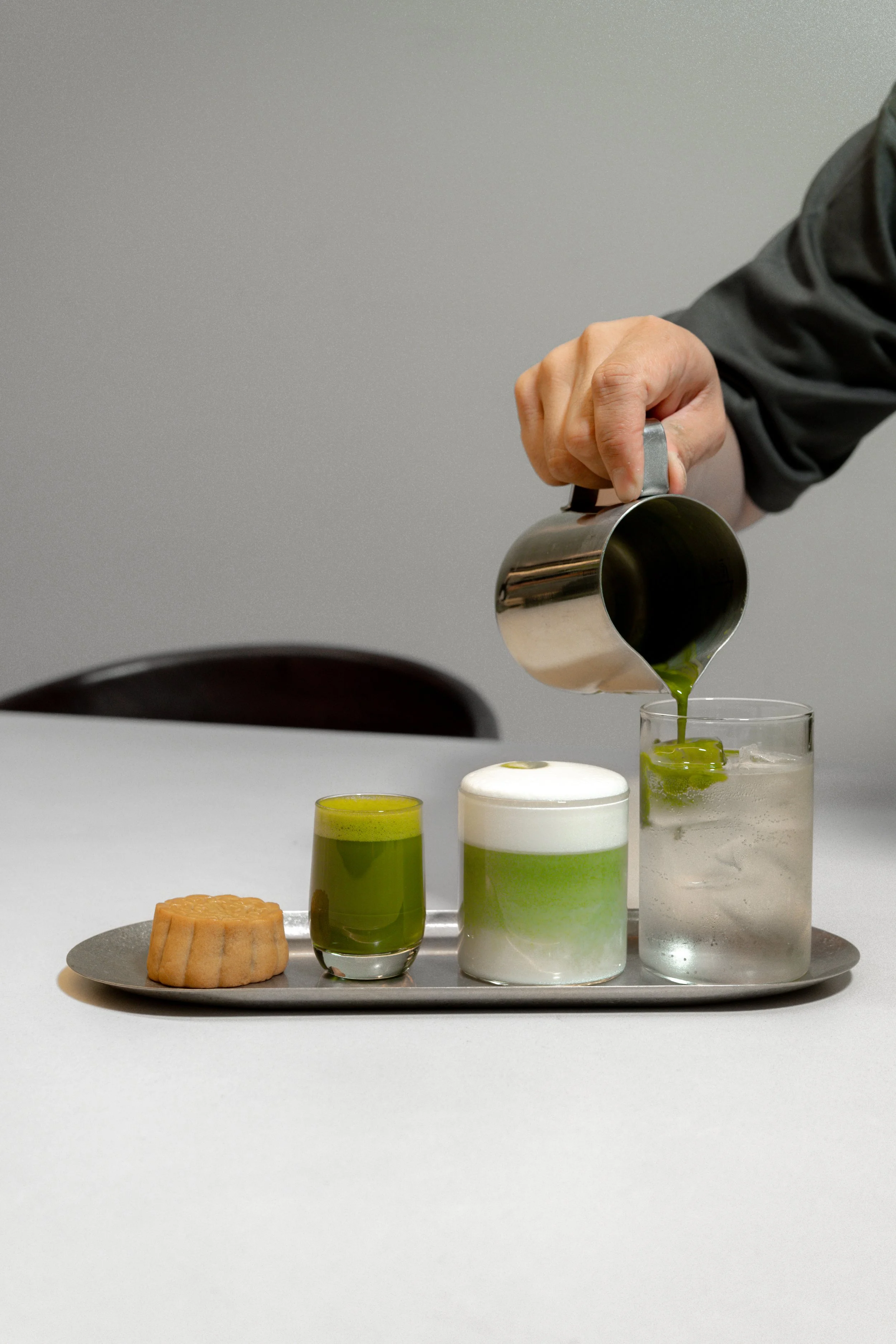 Person pouring matcha in a matcha tonic, with a small matcha latte, a usucha and a mooncake on a tray on a white table.