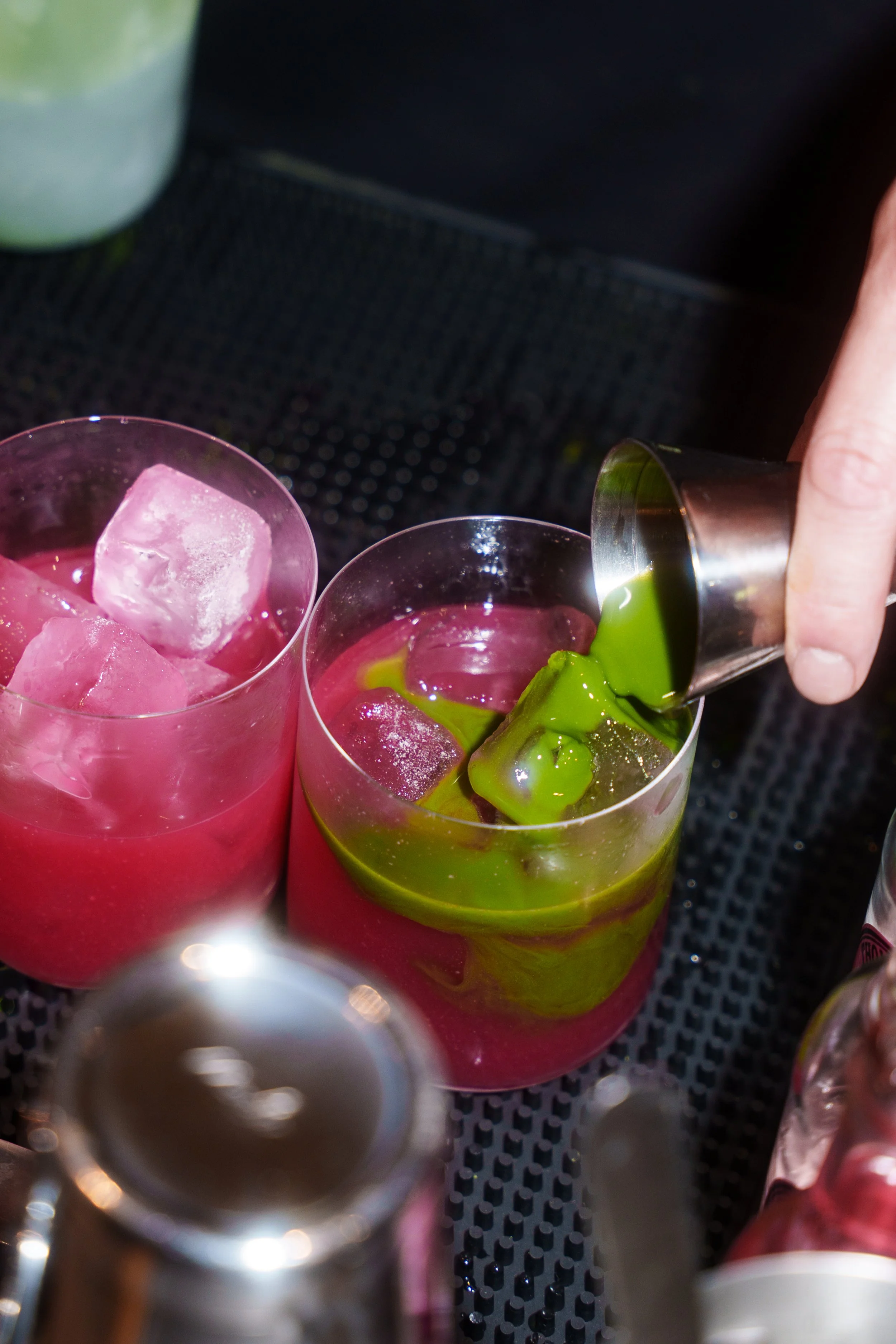 A bartender pouring matcha over pink base with ice in glasses on a bar counter.