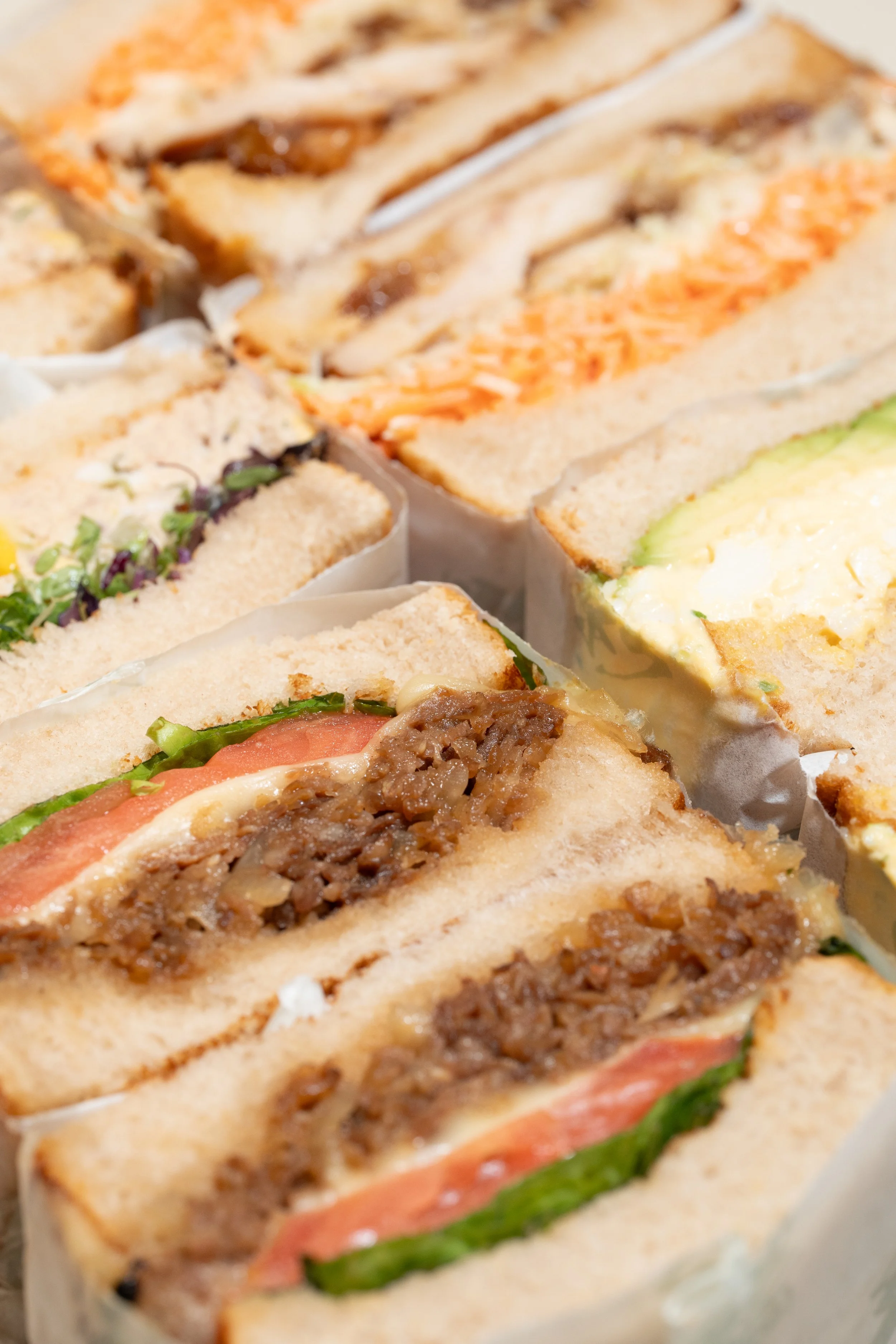 Close-up of various japanese sandwiches (sandos), including one with bulgogi beef, tomato, and lettuce, and other sandwiches with different fillings.