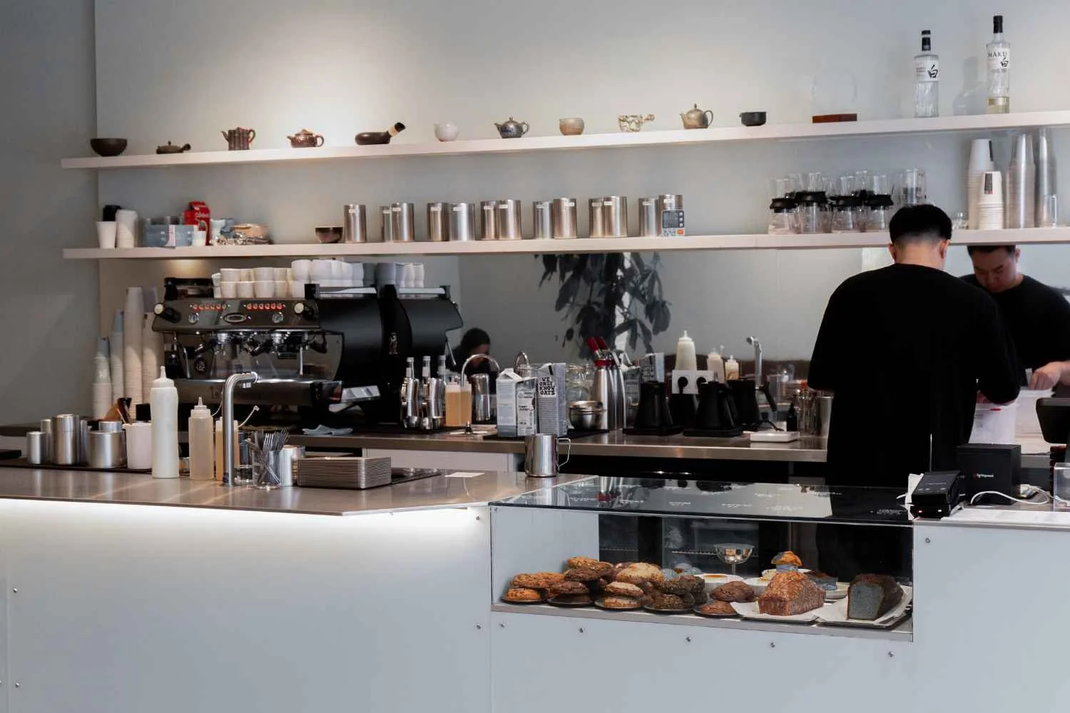 Interior of a matcha and tea bar with one barista preparing drinks behind the counter, shelves with cups, jars, and bottles, and a display of baked goods.