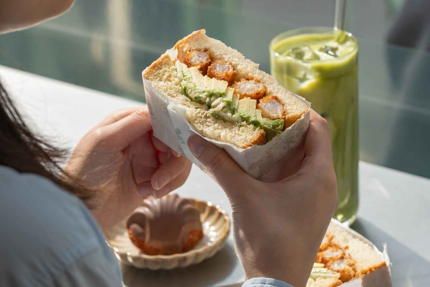 A person holding a japanese sando with shrimp tempura, lettuce, and avocado inside. There is an iced matcha latte, chocolate madeleine on the table.