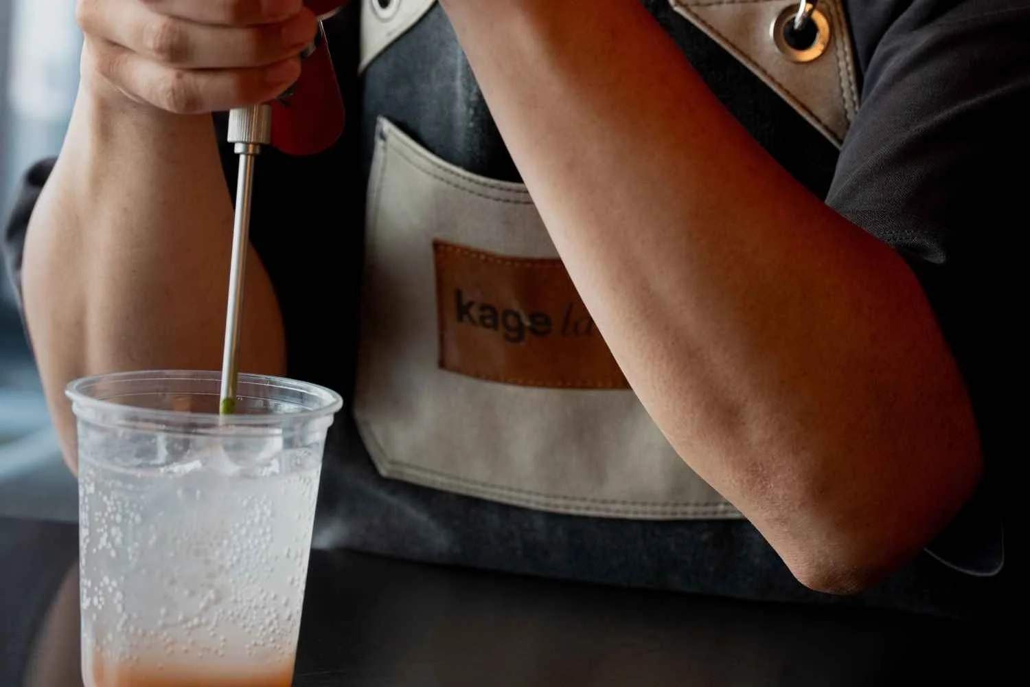 A person is using a tool to stir a clear plastic cup of soda or sparkling beverage, with bubbles visible in the drink. The individual is wearing a black shirt and a gray apron with a brown leather patch labeled 'kagel' and a pocket. The person's arm and hand are visible, holding a stir stick.