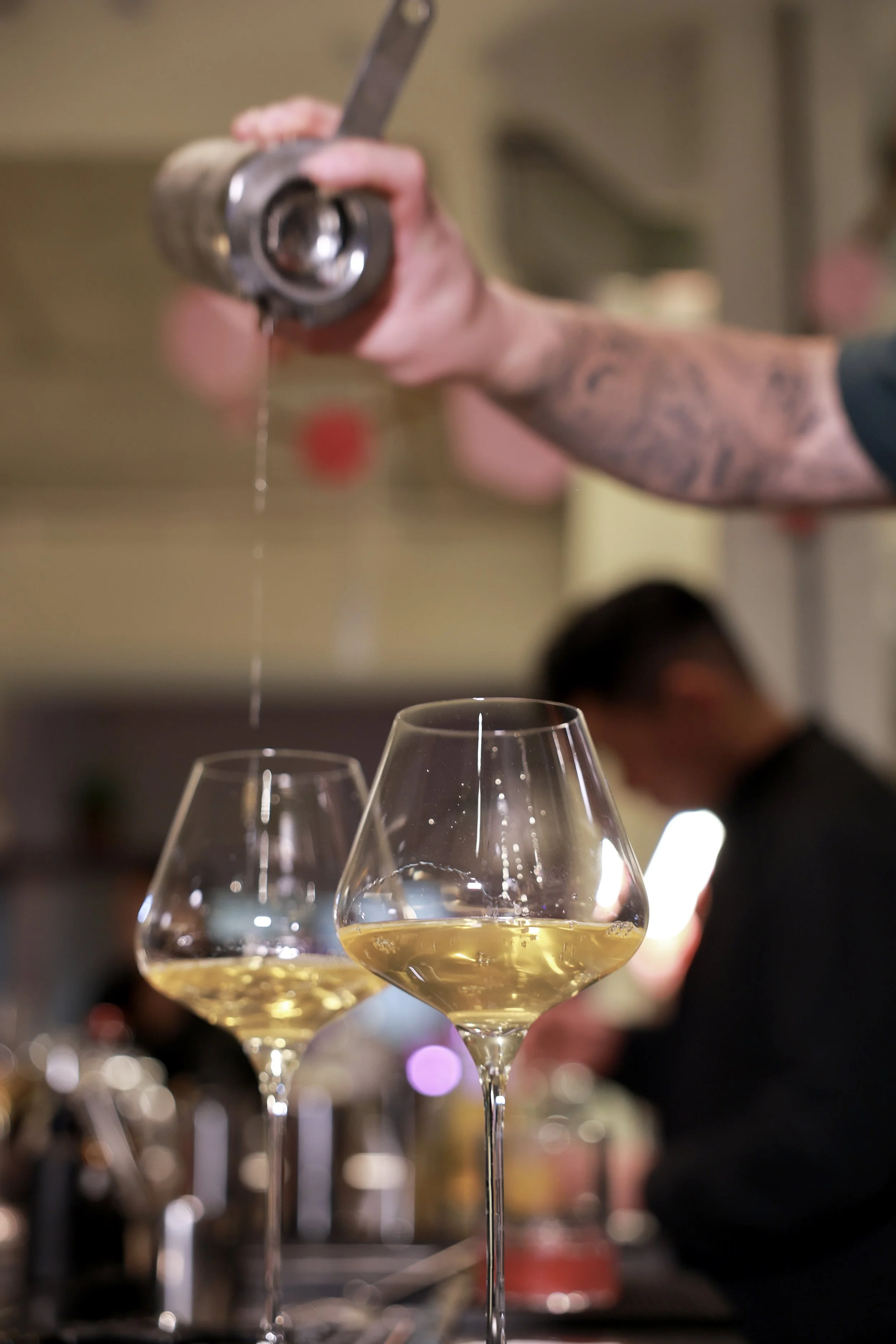 A bartender pouring a tea cocktails drink into wine glasses with a blurred background of people and bar setting.