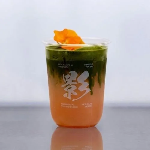 A layered drink with matcha and peach juice topped with an orange flower in a clear glass