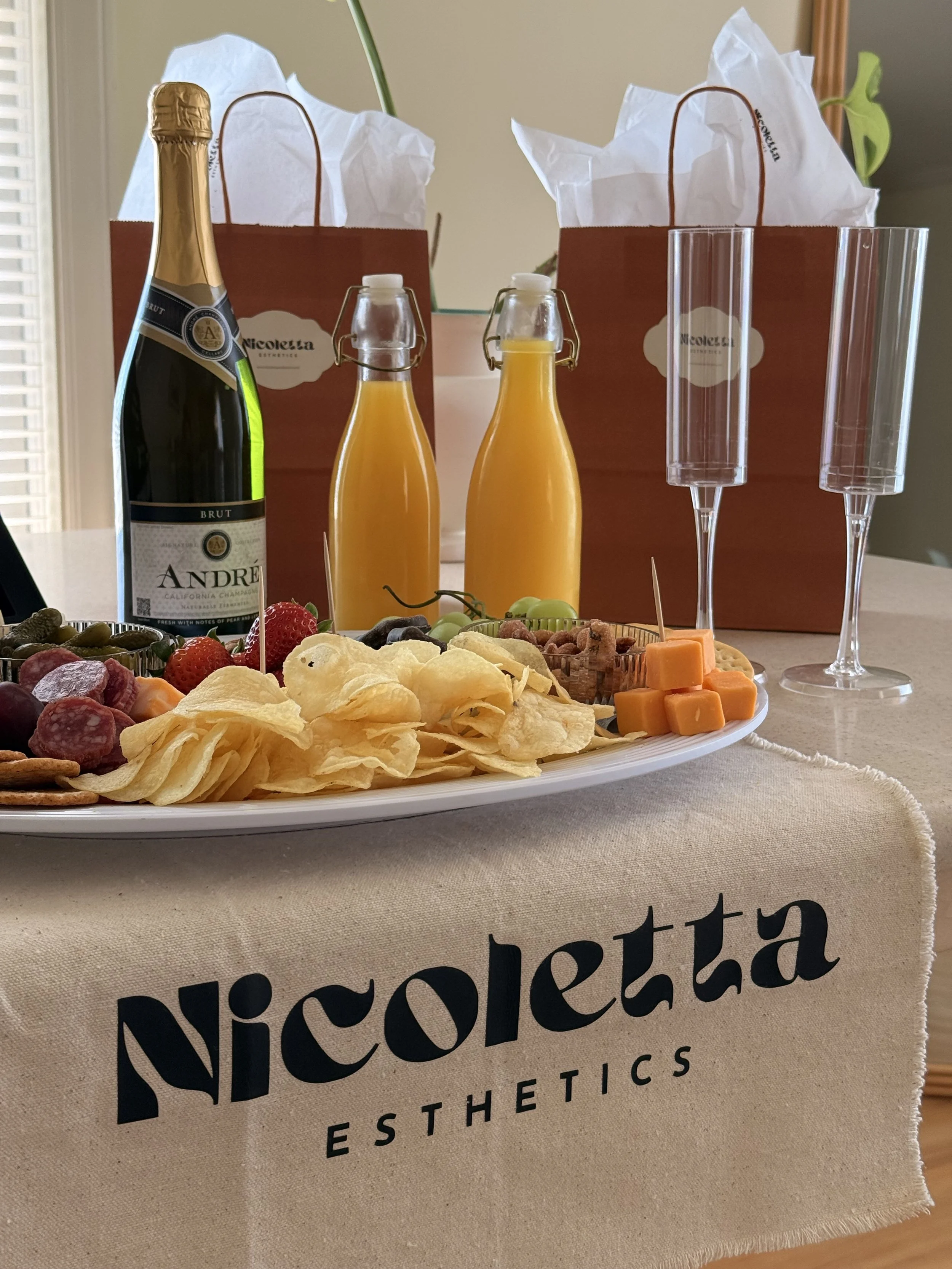 A table with a plate of assorted cheese, grapes, strawberries, crackers, and potato chips. Two bottles of orange juice, a bottle of sparkling wine, and two empty champagne glasses are also on the table. There are two brown gift bags with white tissue paper in the background.
