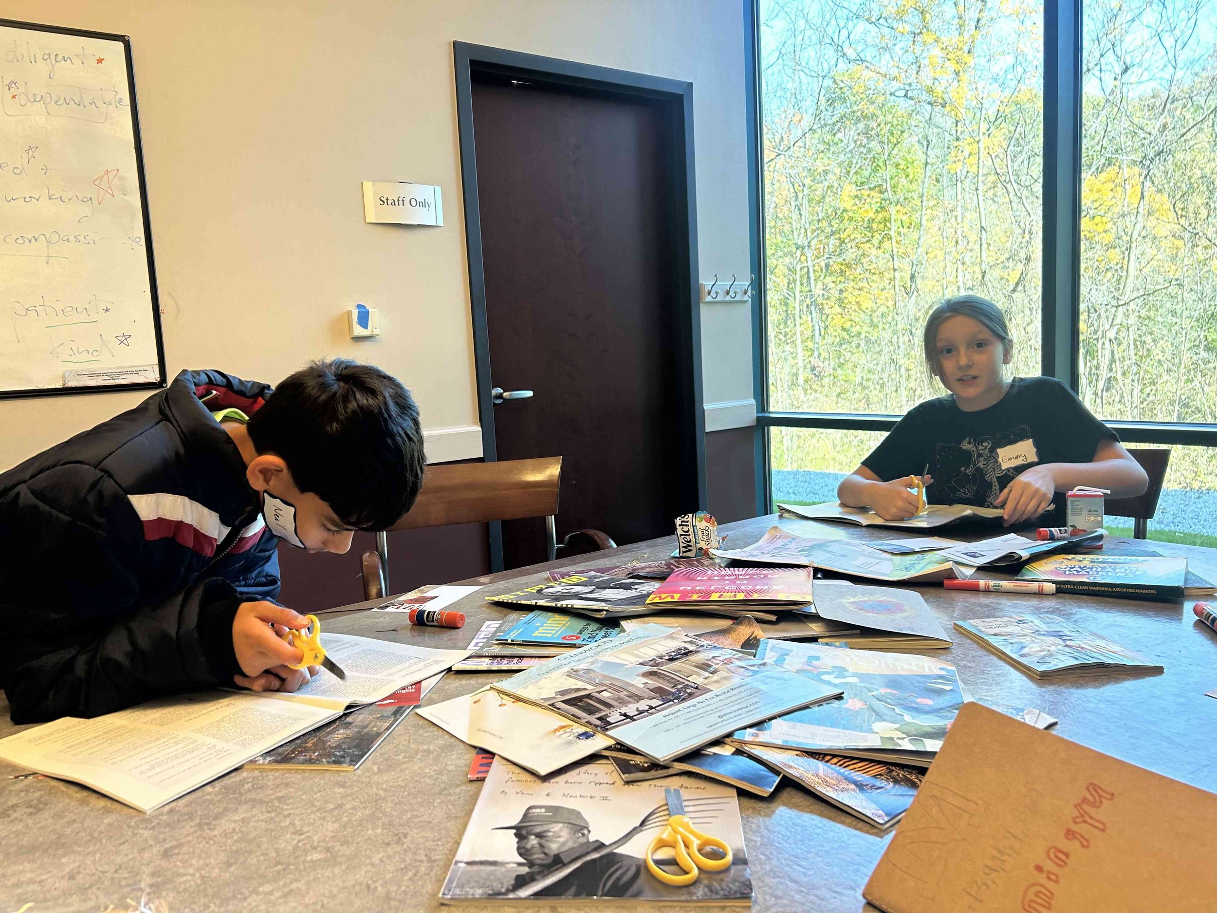 Two children sitting at a table in a room with a window, working on arts and crafts with magazines, scissors, glue, and markers scattered on the table.