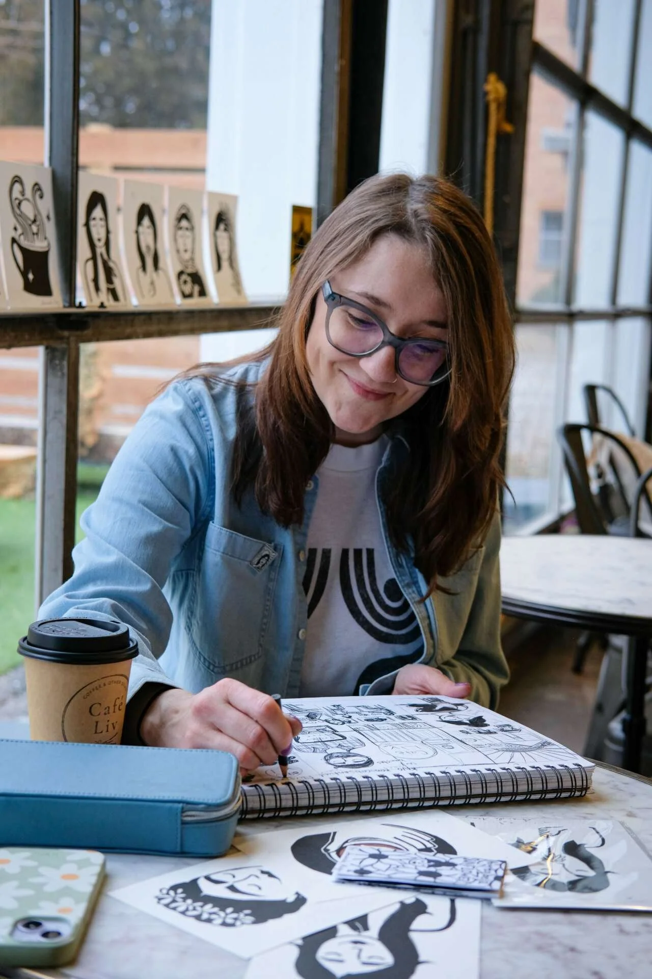 A photo of Amanda May Moore smiling in front of black and white artwork with portrait sketches on a wall.