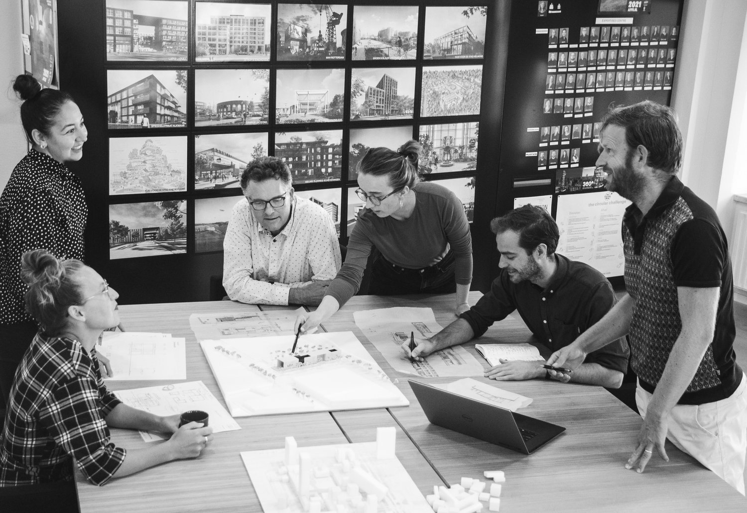 Vijf mensen in een vergaderruimte bekijken en bespreken een model of ontwerp op een tafel, met een achtergrond van meerdere schermen met vastgoedprojecten en plannen.