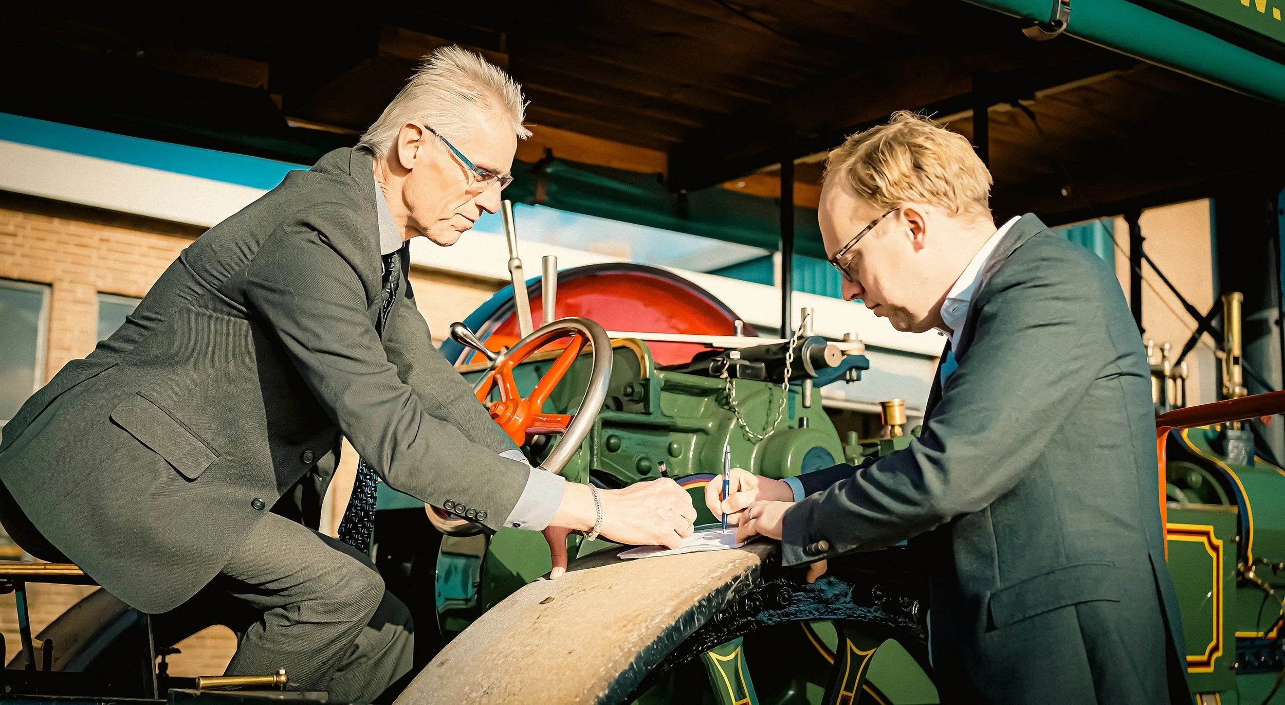 Twee mannen in formele kleding die onder een houten plafond werken aan een groene machine, mogelijk een oude locomotief, terwijl ze documenten en pennen gebruiken.