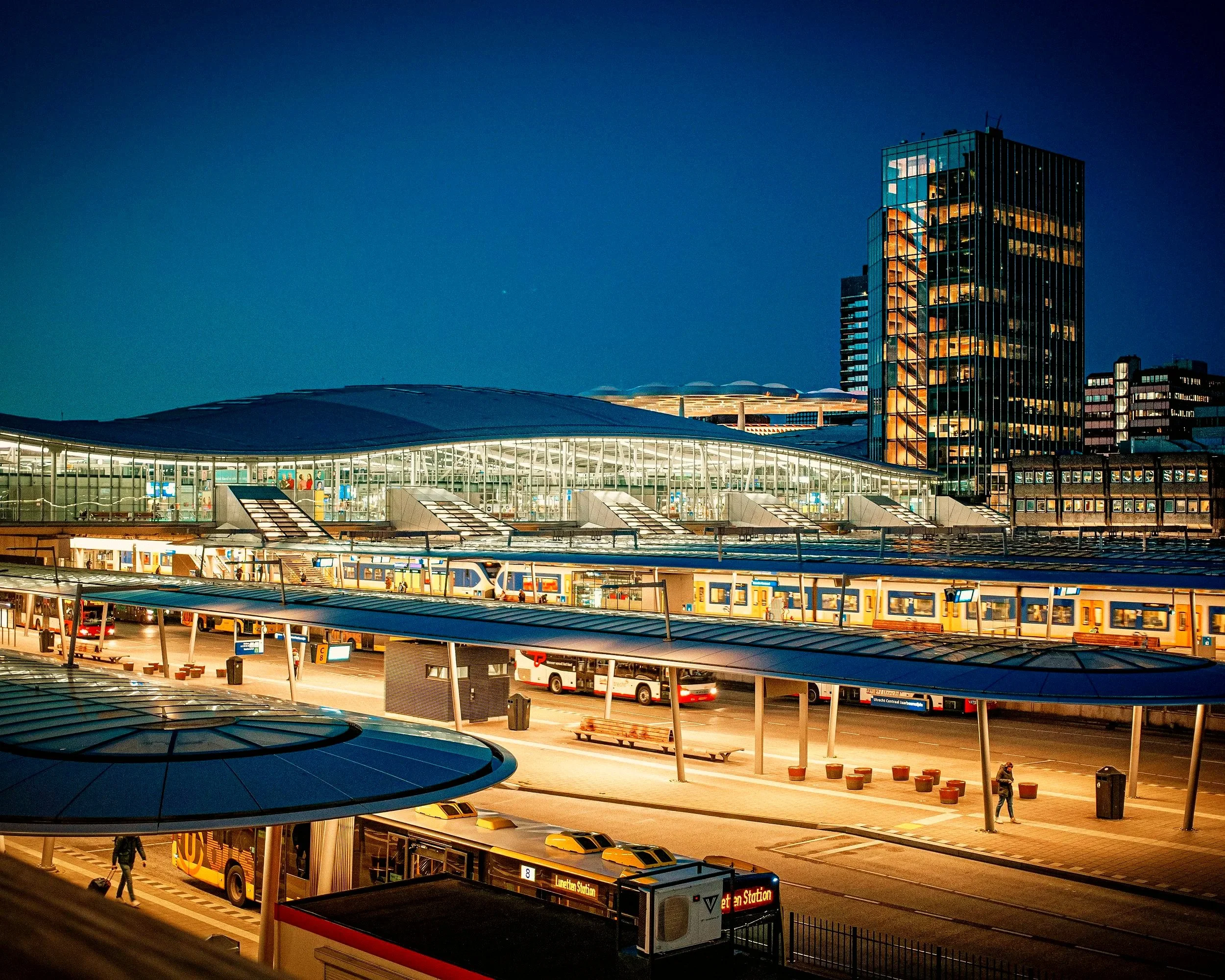 Volgens de foto is het een stadsbeeld 's avonds met een modern kantoorgebouw met grote glazen ramen en een hoog gebouw daarnaast. In de onderliggende laag is een treinstation met een paar bussen en auto's te zien, en enkele mensen lopen rond.