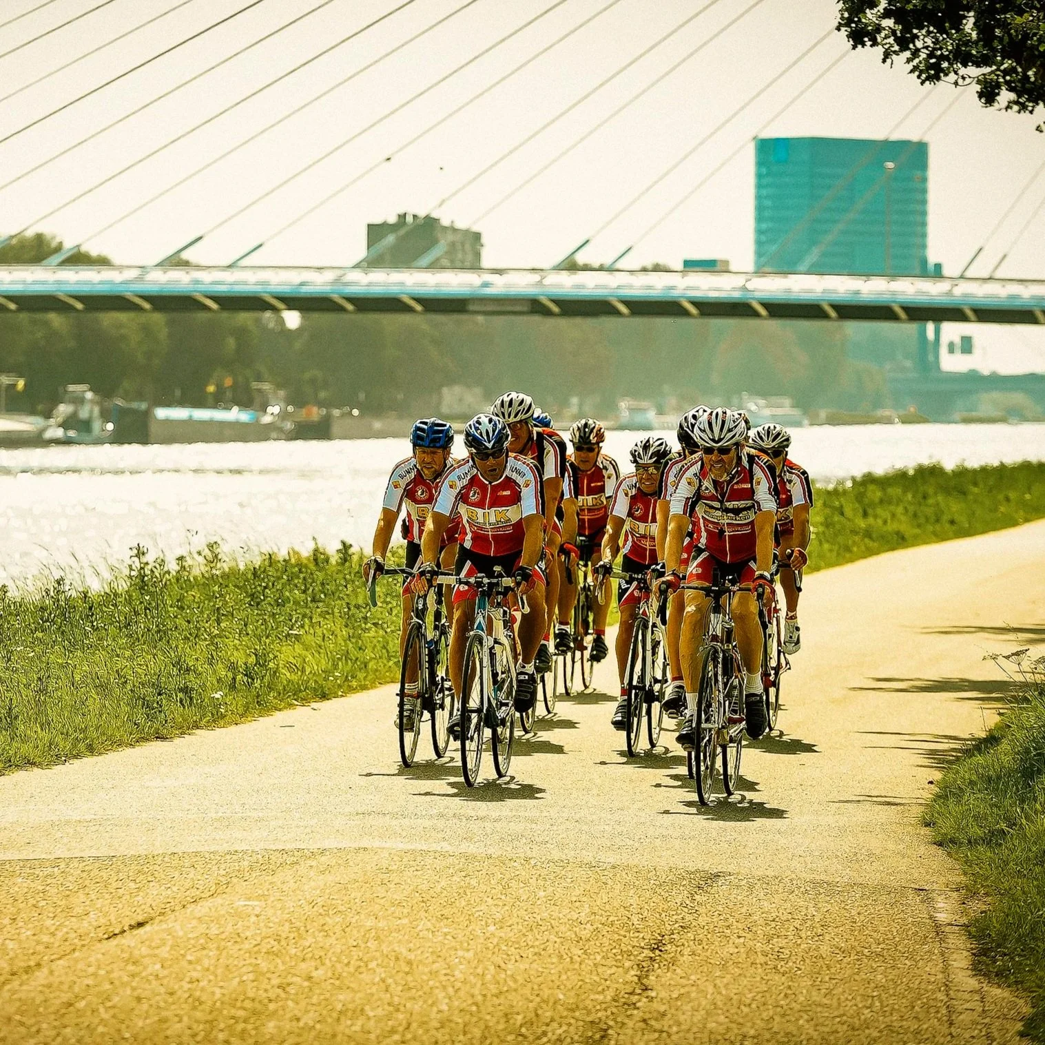 Een groep wielrenners fietst samen op een zonnige dag langs een rivier met een brug op de achtergrond.
