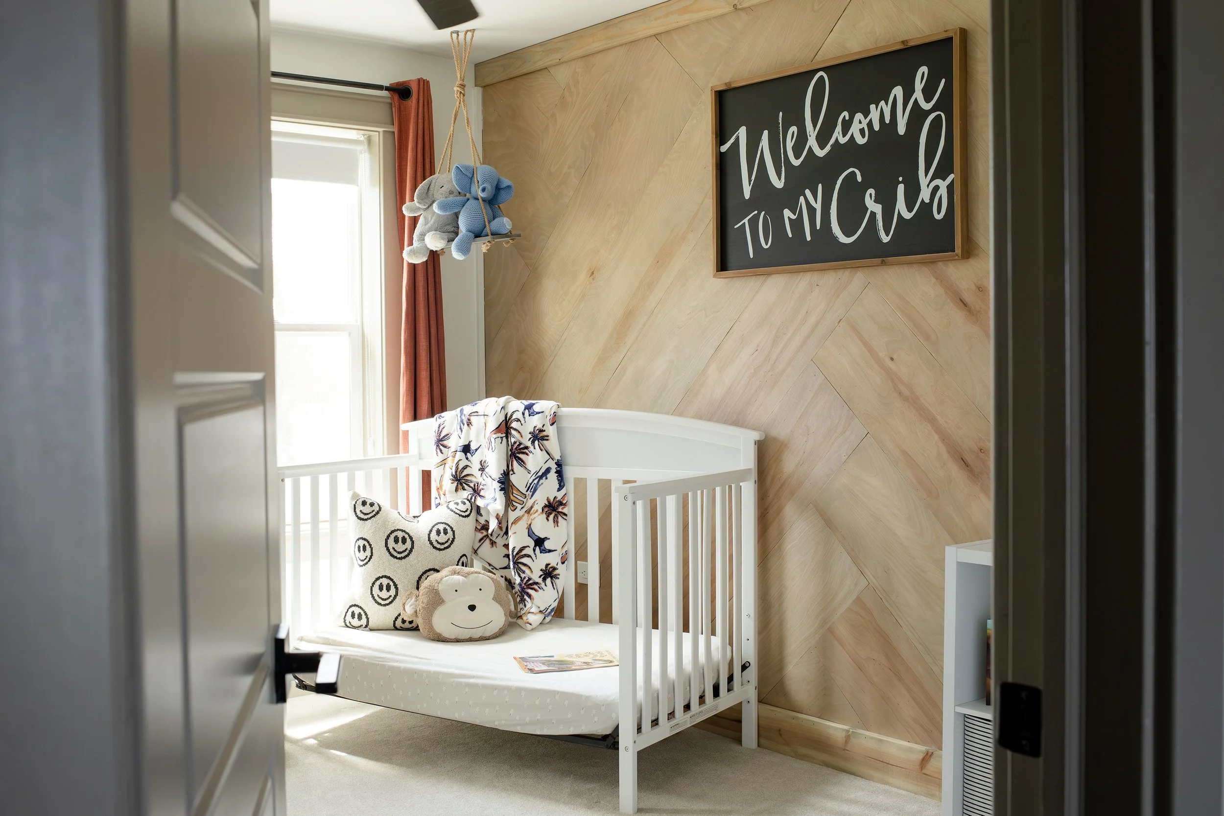 Nursery room with a white crib, plush monkey and pillow, a blanket, a window with orange curtains, a blackboard sign that reads 'Welcome to my crib', and a mobile with stuffed animals hanging from the ceiling.