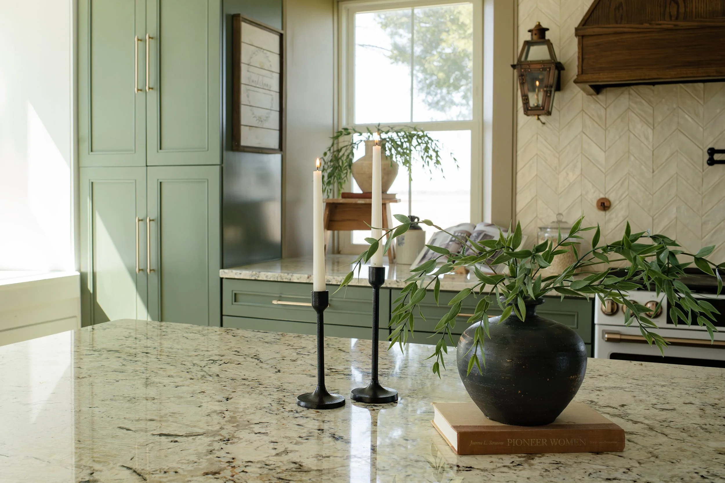 A kitchen with green cabinets, a granite countertop, a black vase with green leafy branches, two black candle holders with burning candles, a window with trees outside, and a beige textured backsplash.