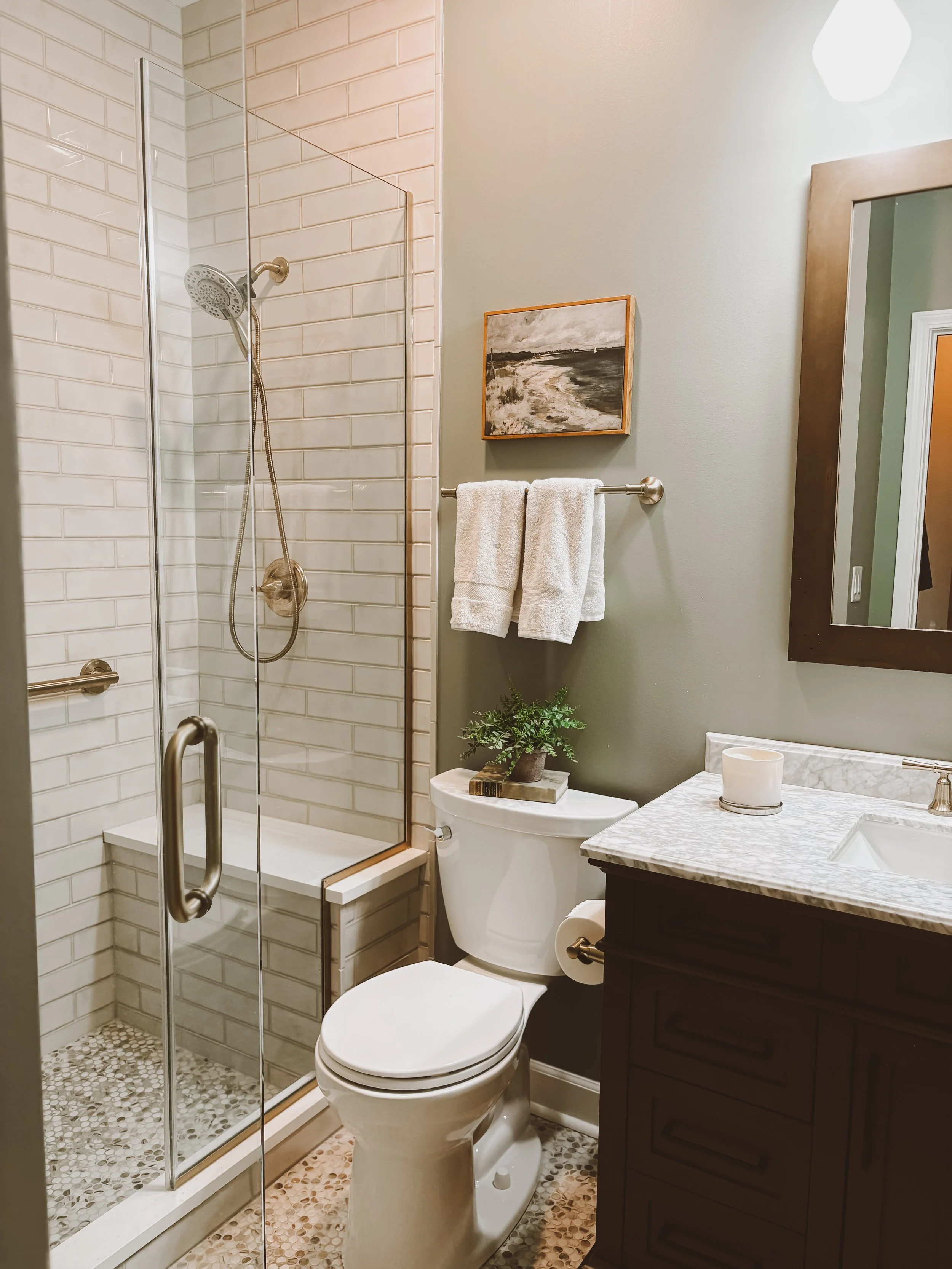 Bathroom with a glass shower enclosure, a white toilet with a potted plant on top, a dark wooden vanity with a white marble countertop, a mirror, a towel rack with white towels, and a wall painting. The floor has small pebble tiles.