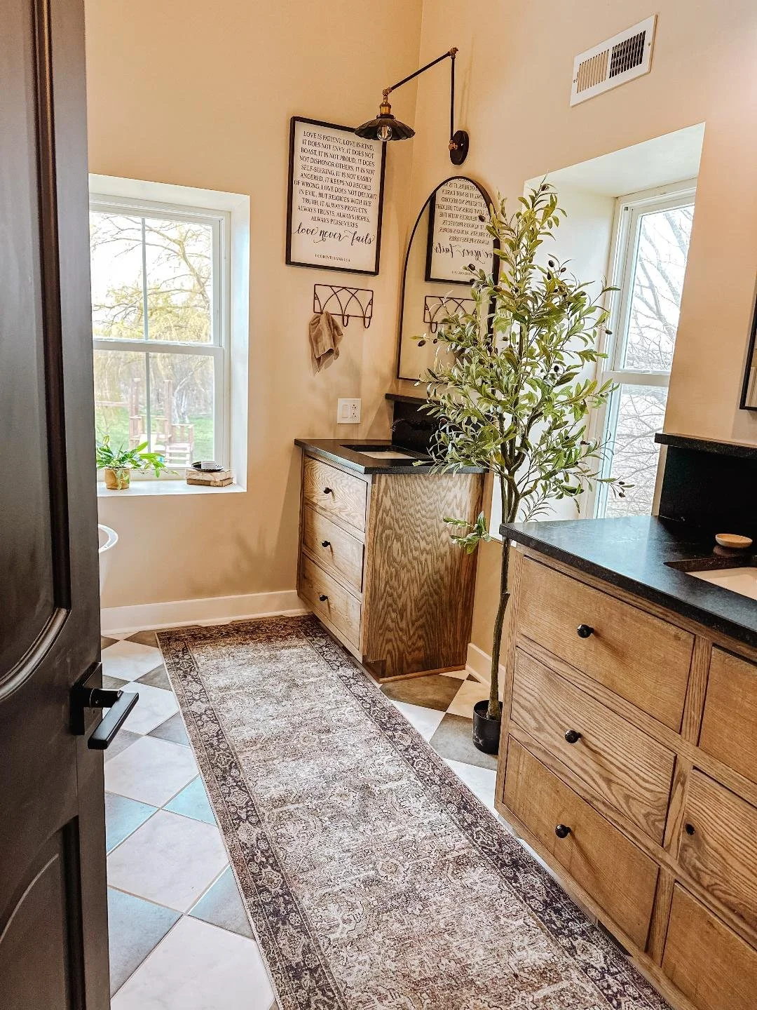 A cozy kitchen corner with two windows, wooden cabinets, a potted plant, framed quotes on the wall, and a patterned rug on tile flooring.