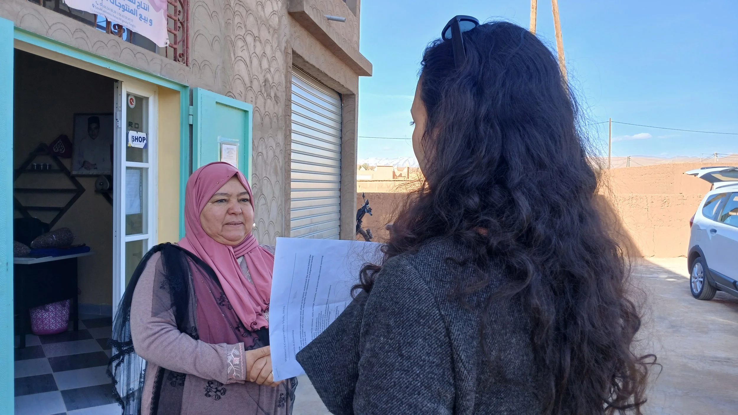 A woman in a gray jacket and sunglasses talking to an older woman wearing a pink hijab and beige sweater outside a building. The older woman is holding a piece of paper, and they appear to be engaged in conversation.