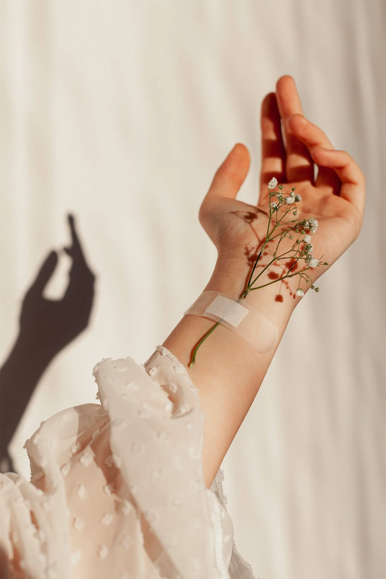 Close-up of a hand with a small flower sprig and a medical bandage, casting a shadow on a white background. The hand is dressed in a white, textured sleeve.