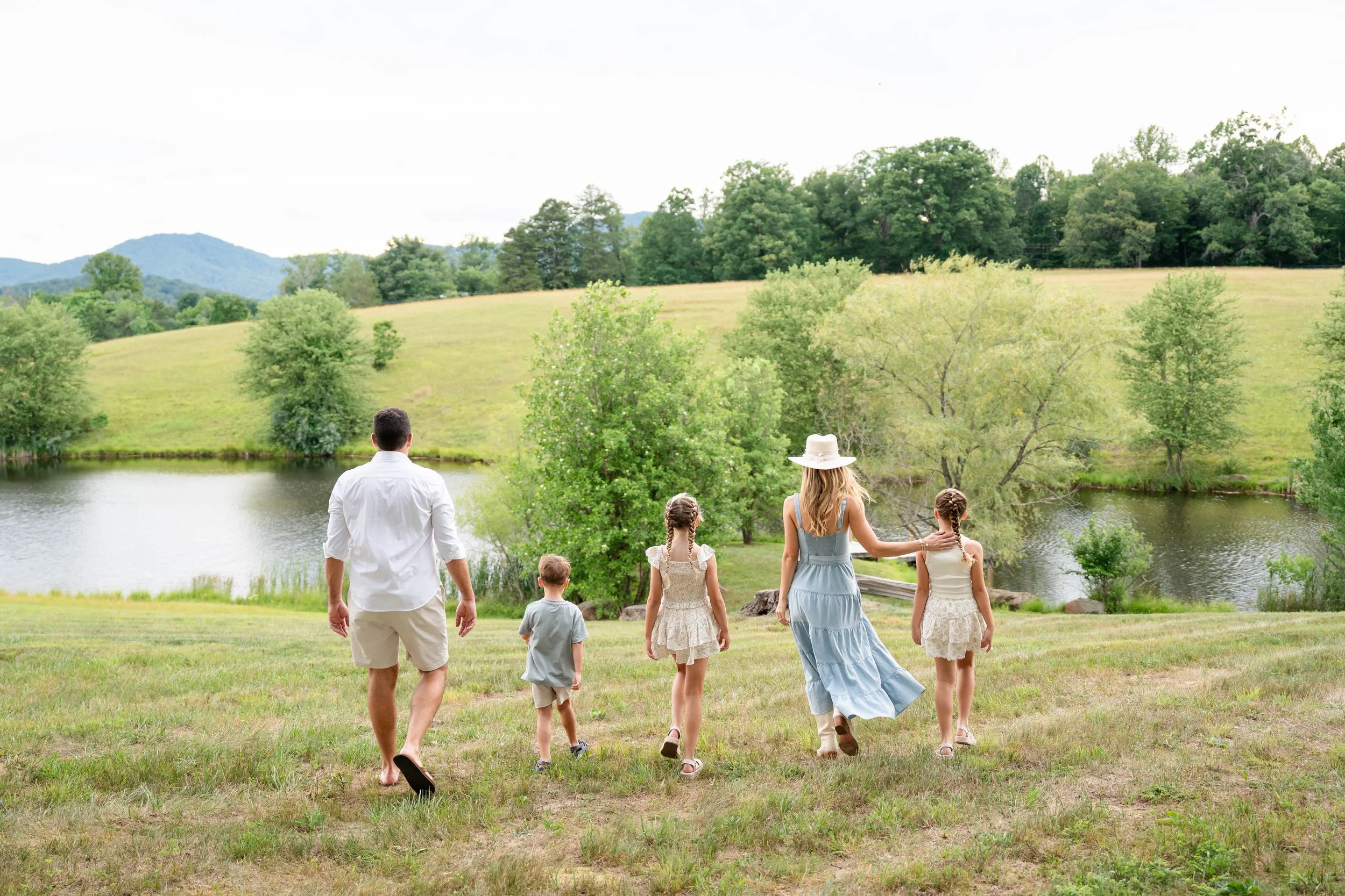  Roehl family walking down a grassy hillside toward the pond at Grey Oaks Farm with the Virginia mountains in the distance. 