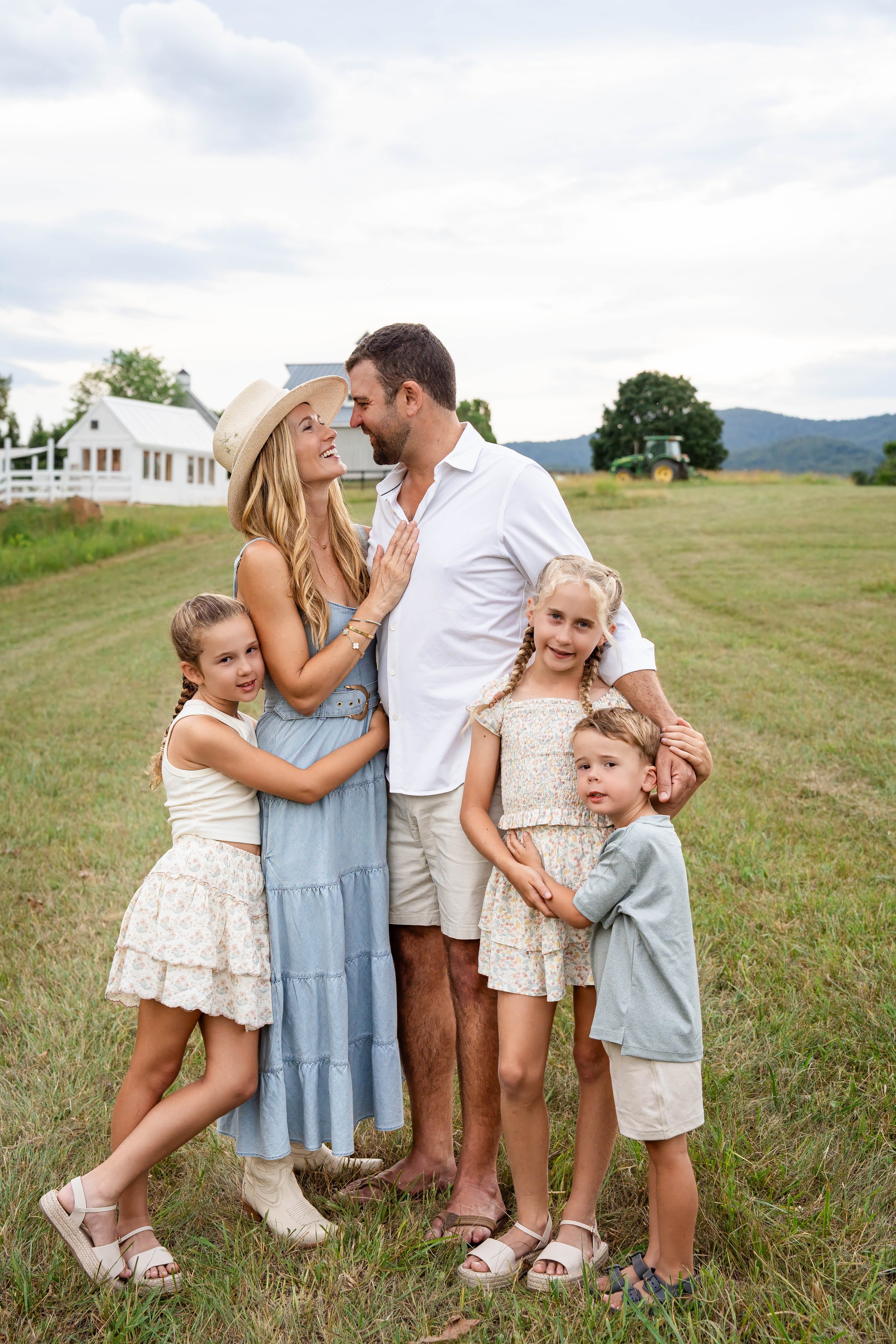 Roehl family of five embracing in a grassy field with farm buildings and Virginia mountain scenery in the background