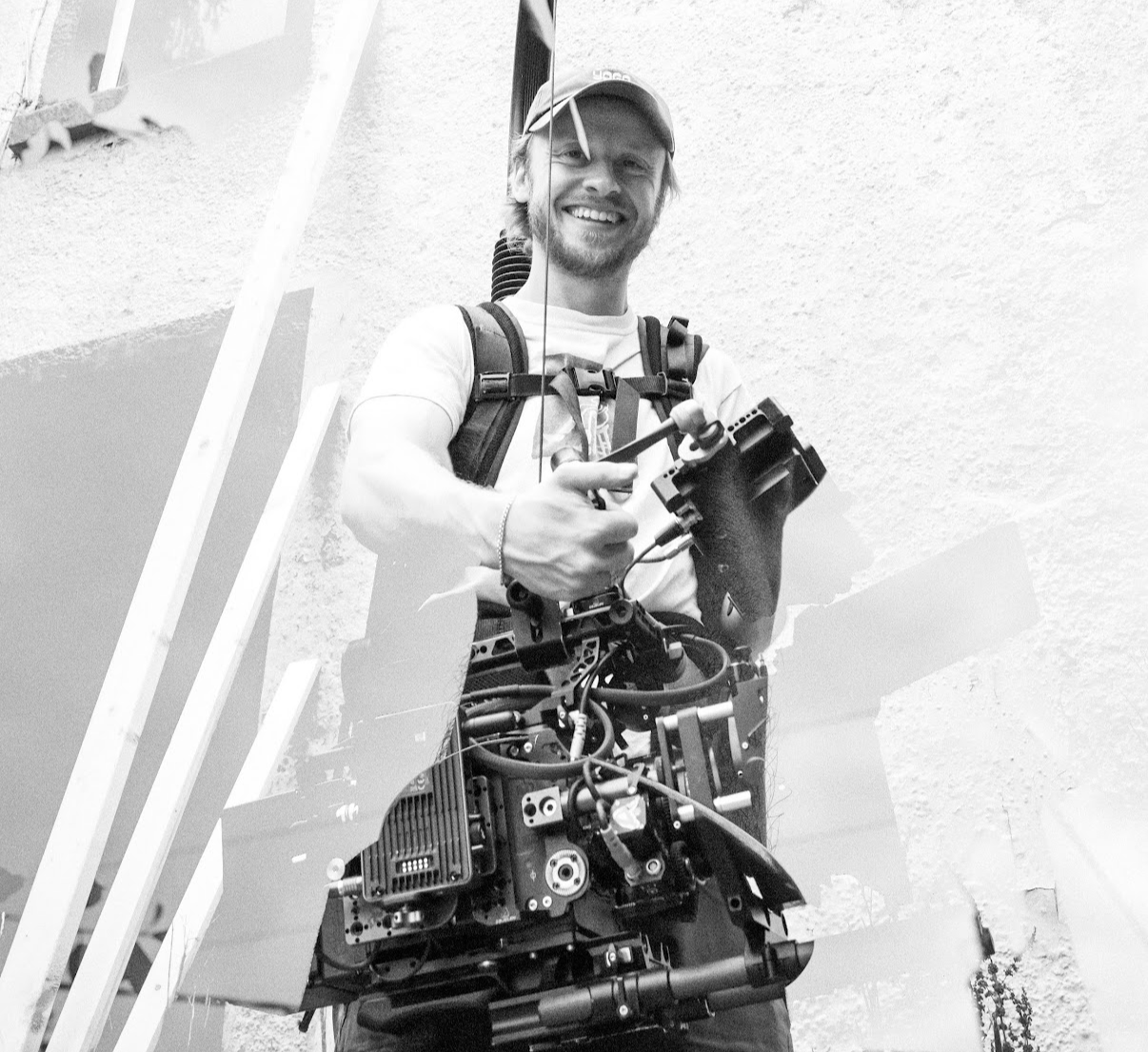 A smiling man wearing a cap and backpack, holding a camera stabilizer, standing in front of a textured wall.